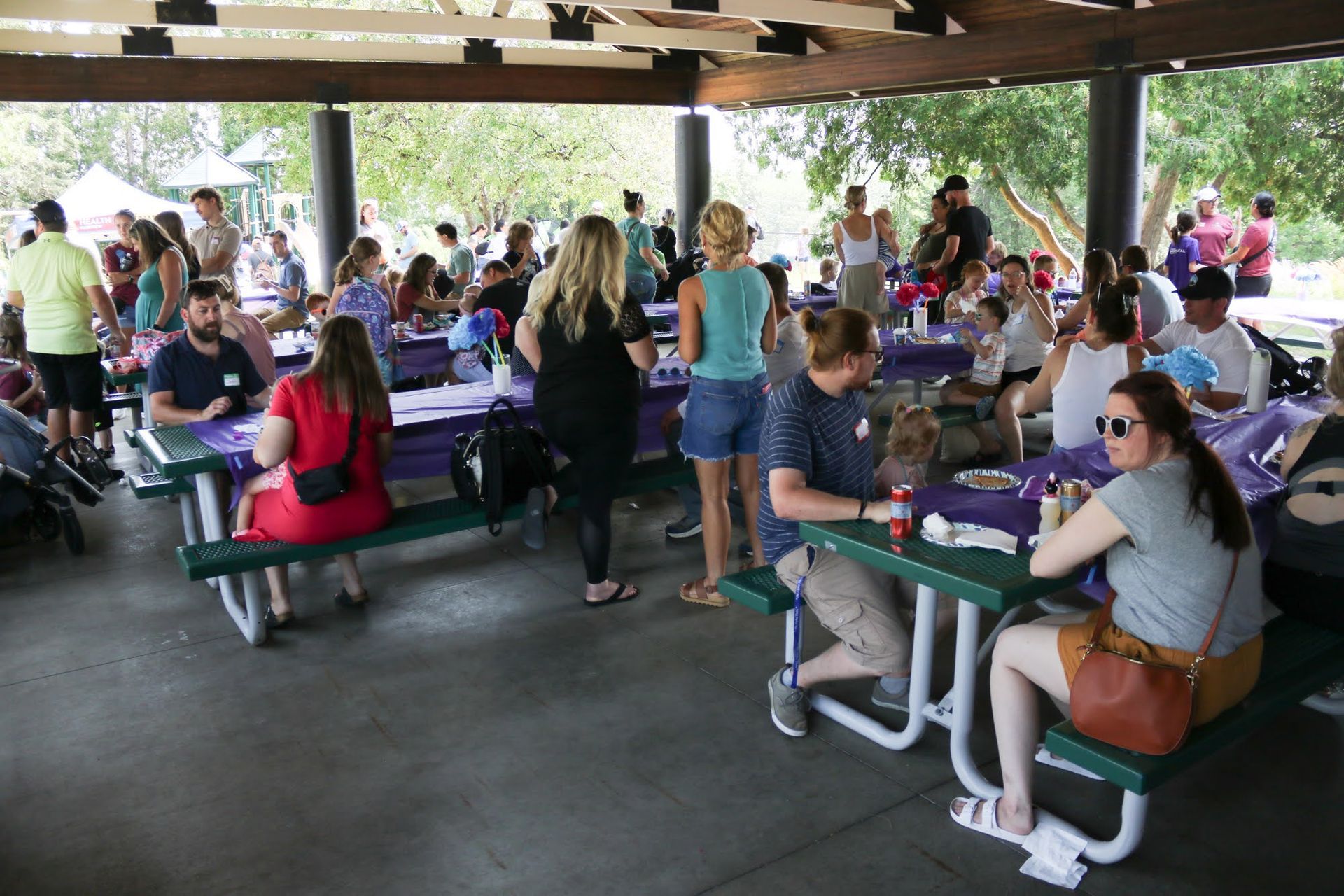People gather at picnic tables under a pavilion for an outdoor event.