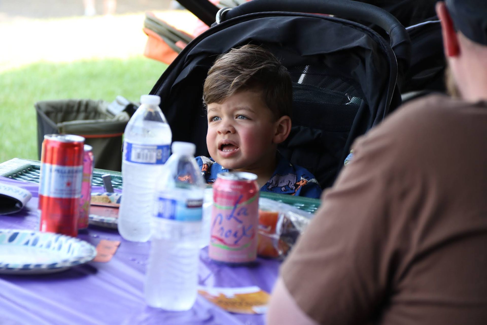 Child in stroller at outdoor table, with an upset expression, surrounded by drinks and a person.