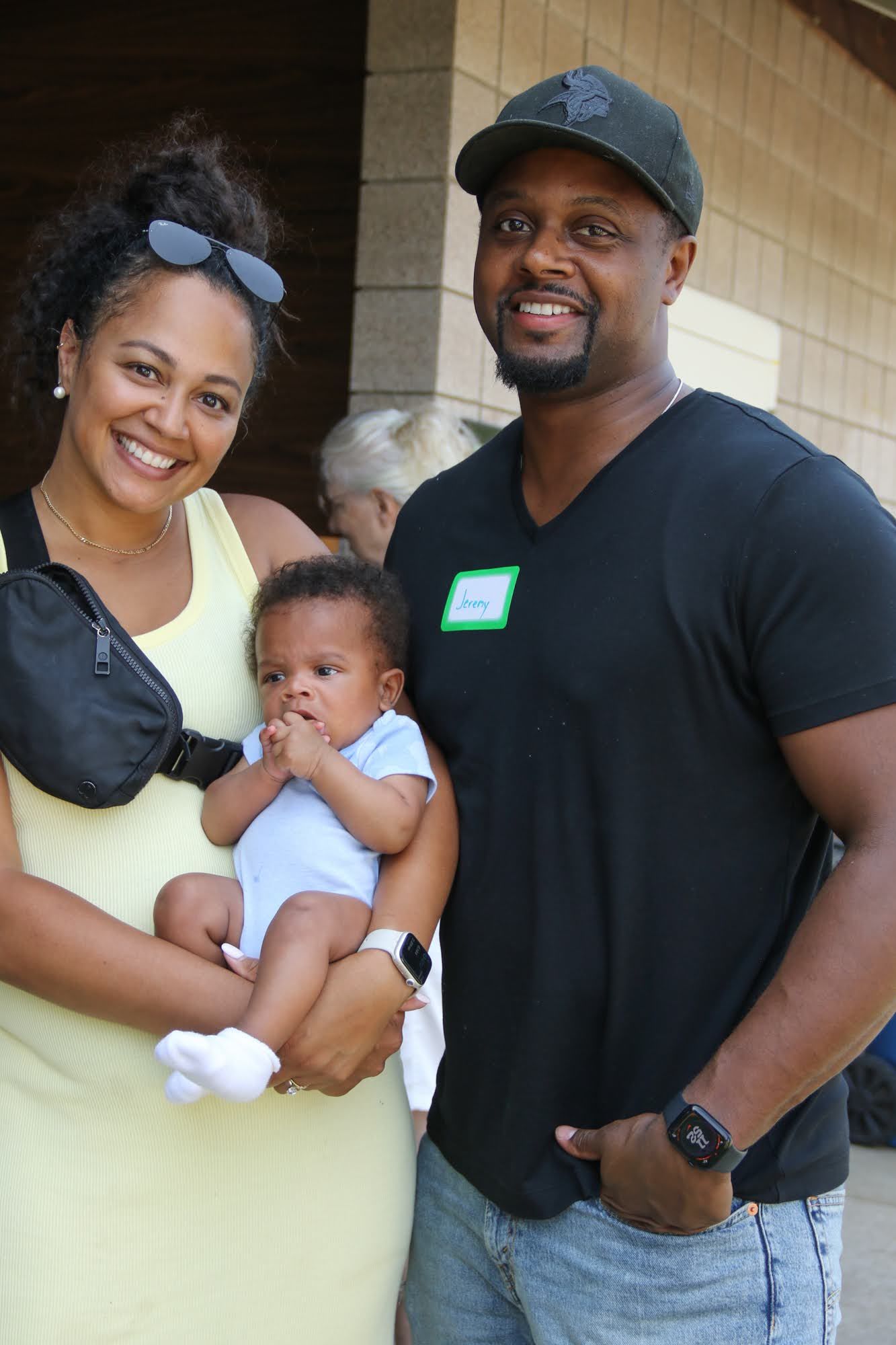 Family of three poses outdoors: woman holds baby, man stands beside them, all smiling.