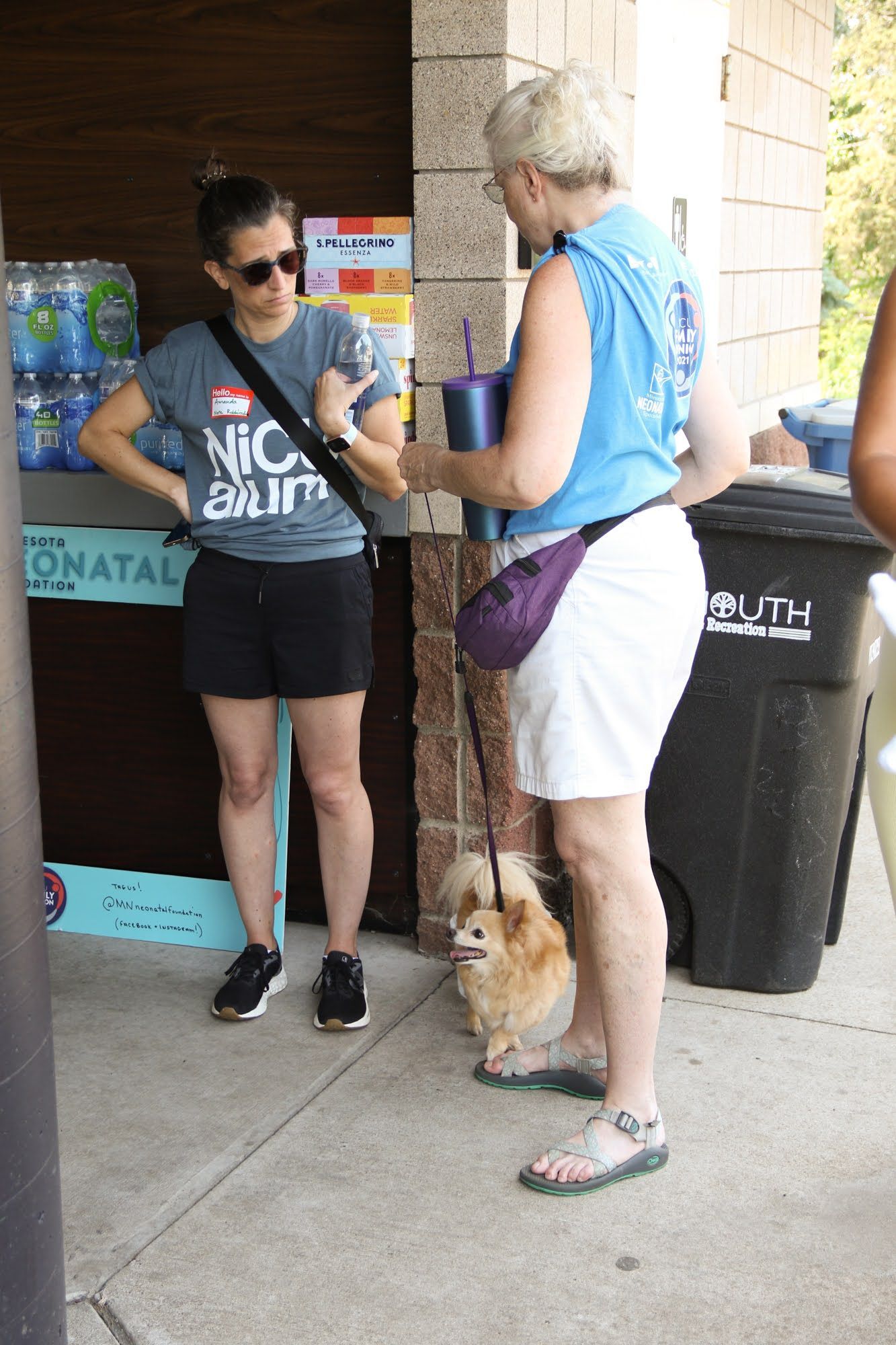 Two people standing, talking near a water bottle display, one with a small dog on a leash.