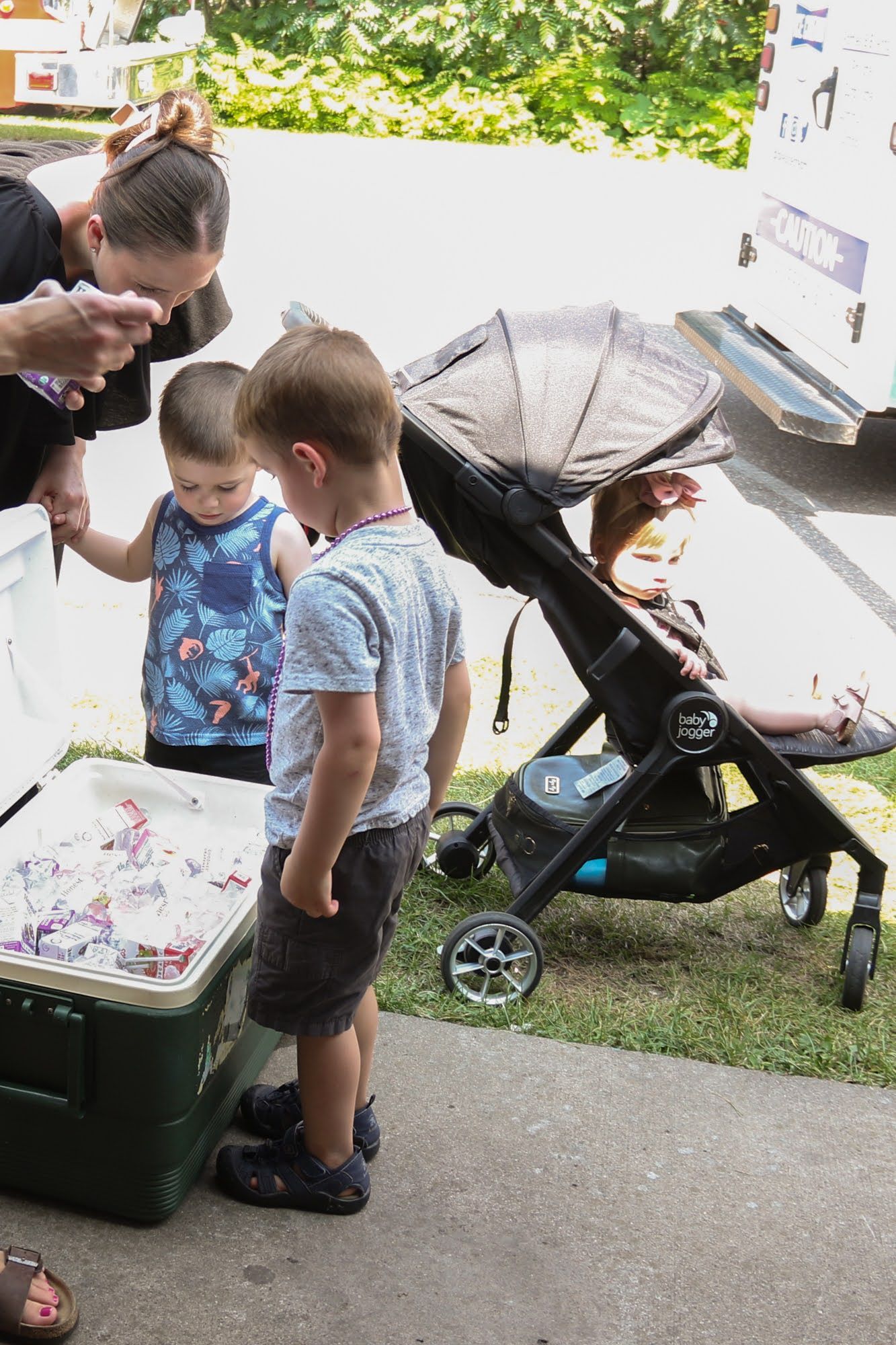 Children look at items in a cooler while a person holds a child's hand. Stroller nearby. Outdoors.