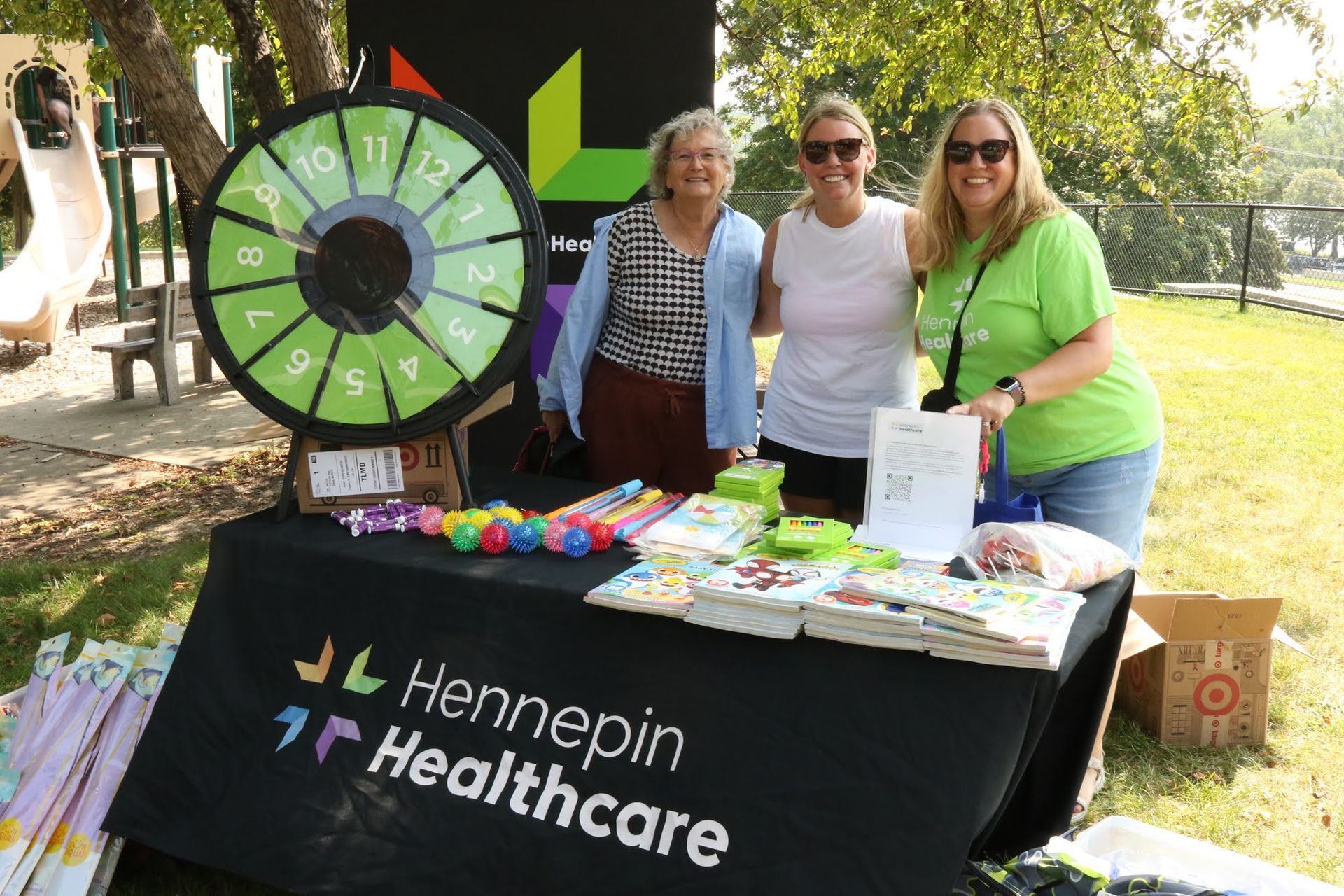 Three women at a Hennepin Healthcare booth outdoors, next to a prize wheel, with giveaway items on the table.