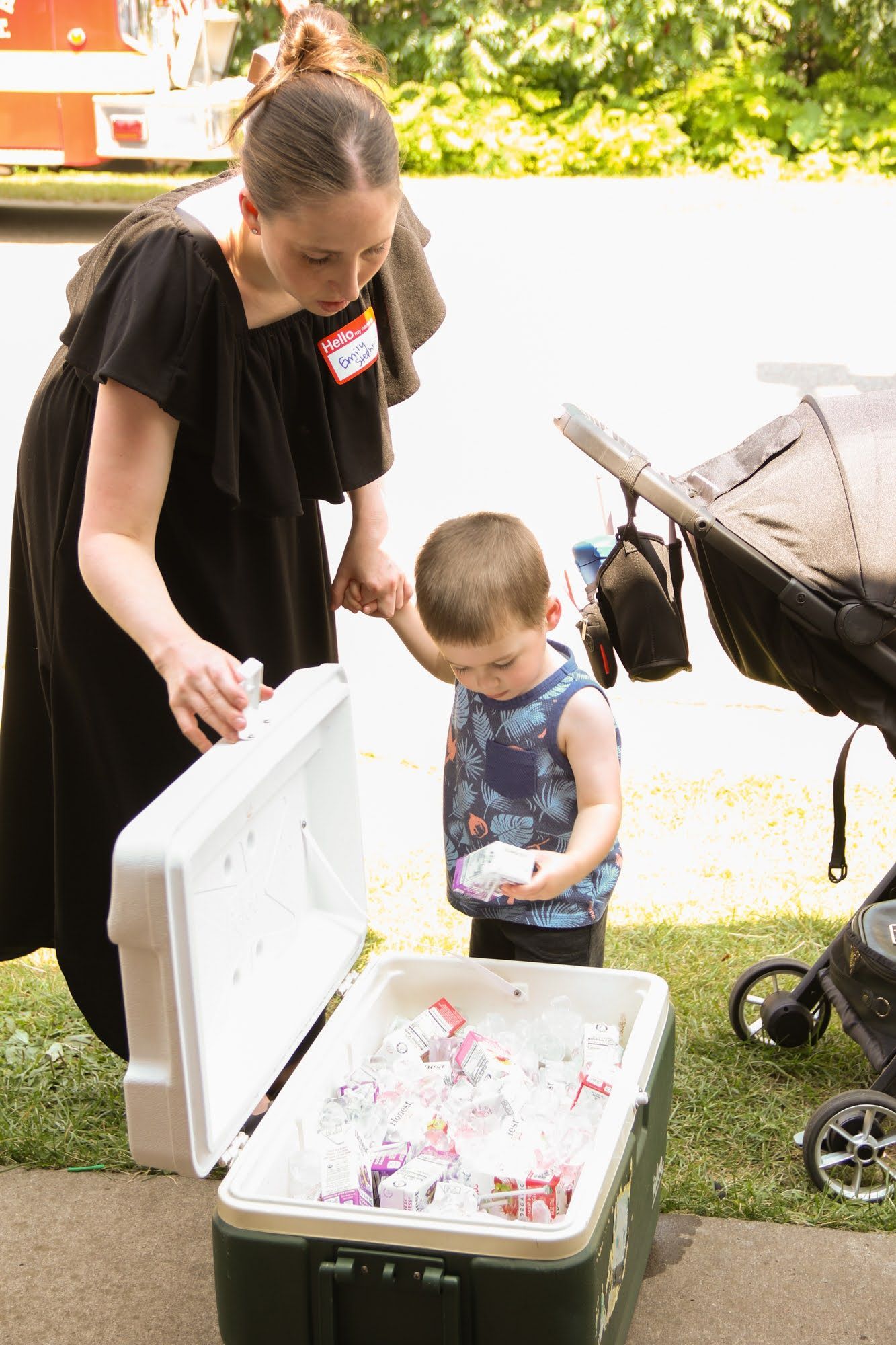 Woman and young child reaching into a cooler filled with drinks. Next to them is a stroller. Outdoors.