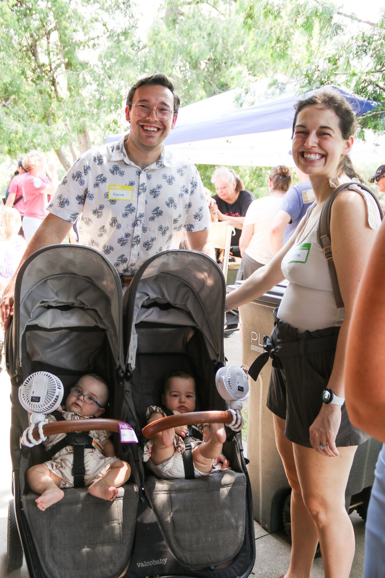 Two adults smile, standing by a double stroller with two babies. Outdoors, trees in background.