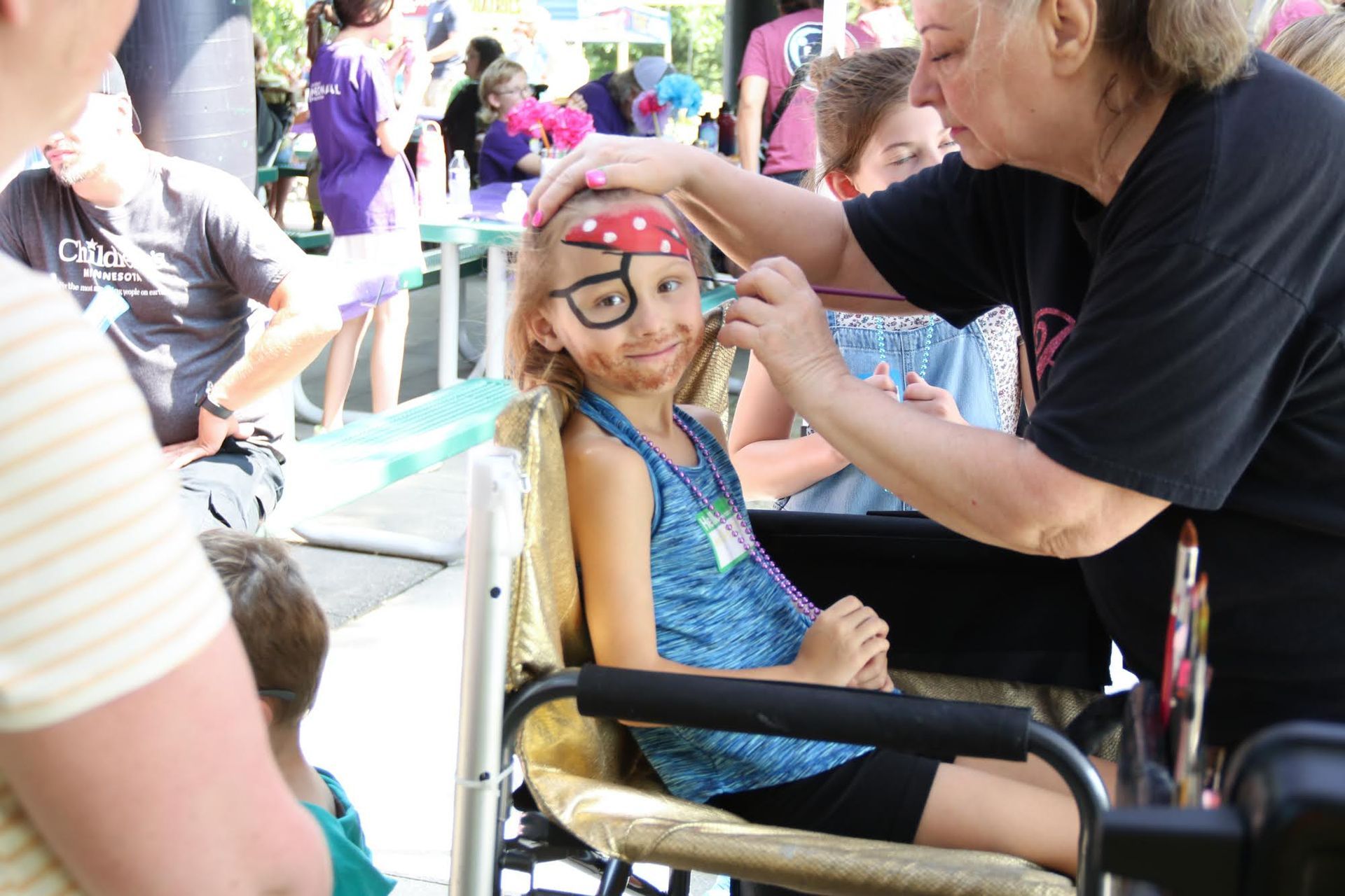 Girl in wheelchair gets pirate face paint at an outdoor event, being applied by a woman.