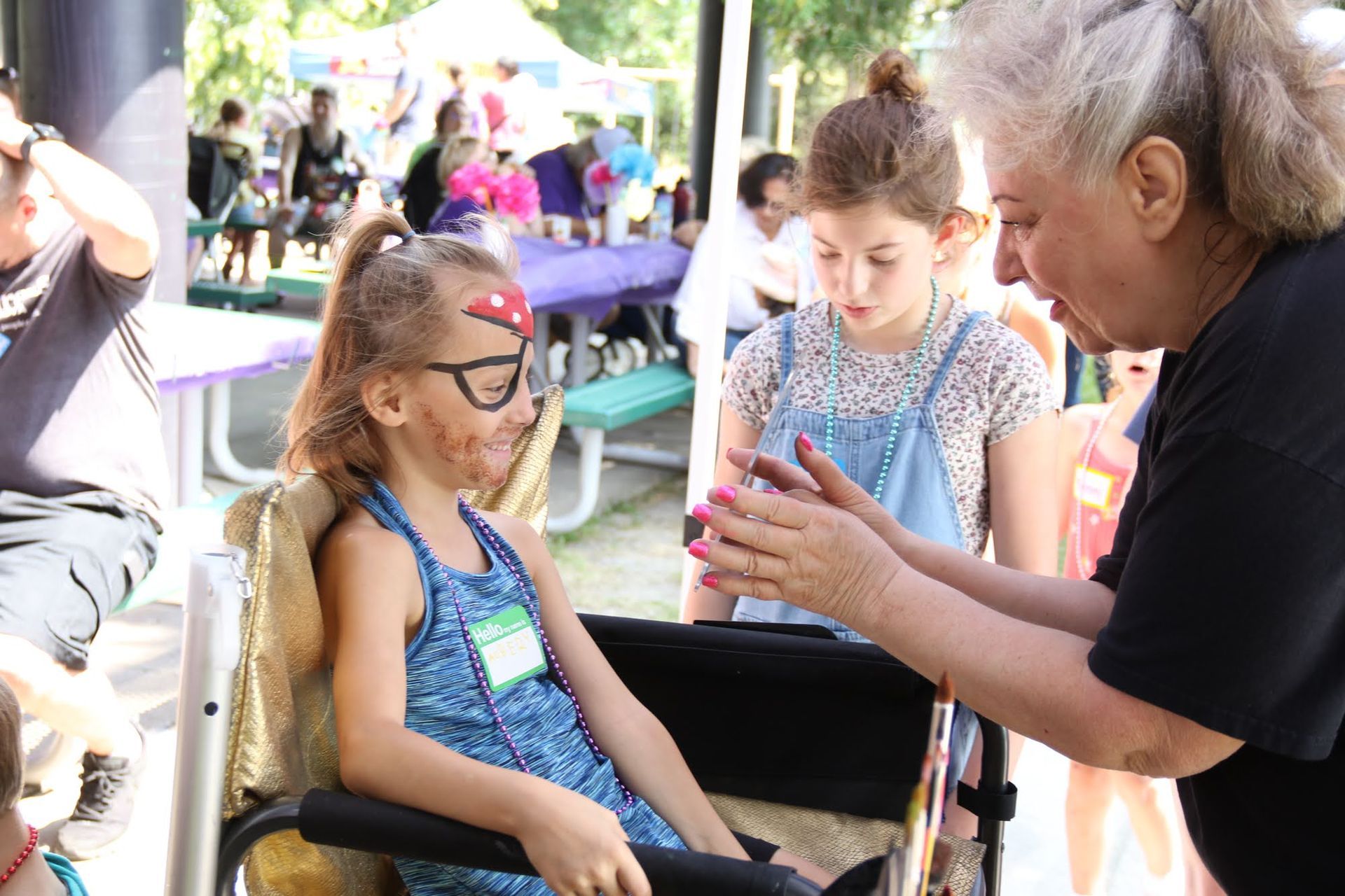 Girl in wheelchair gets face paint at an outdoor event. Another girl and a woman stand nearby.