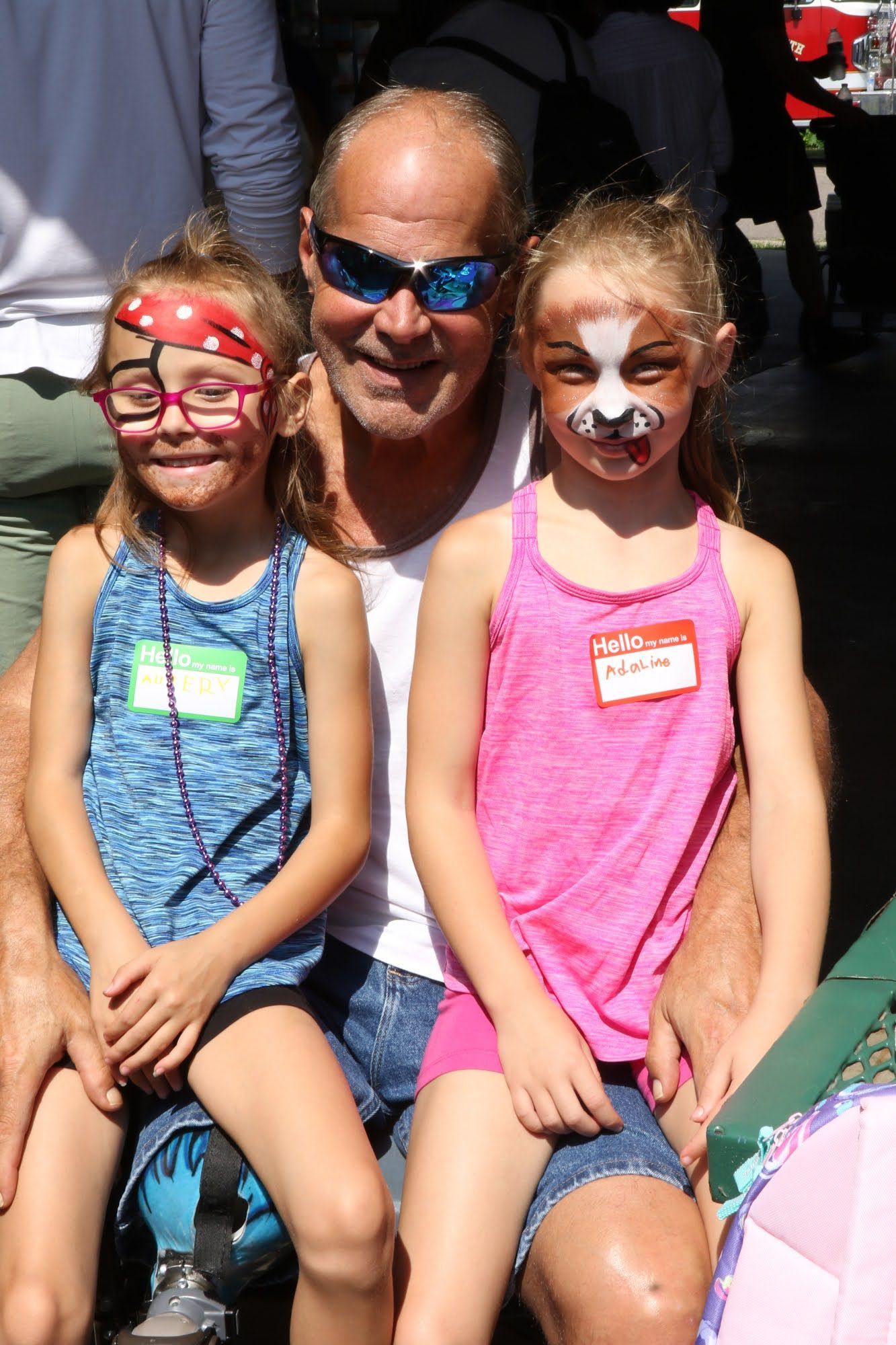Man with two girls, all smiling; faces painted. Outdoor setting.