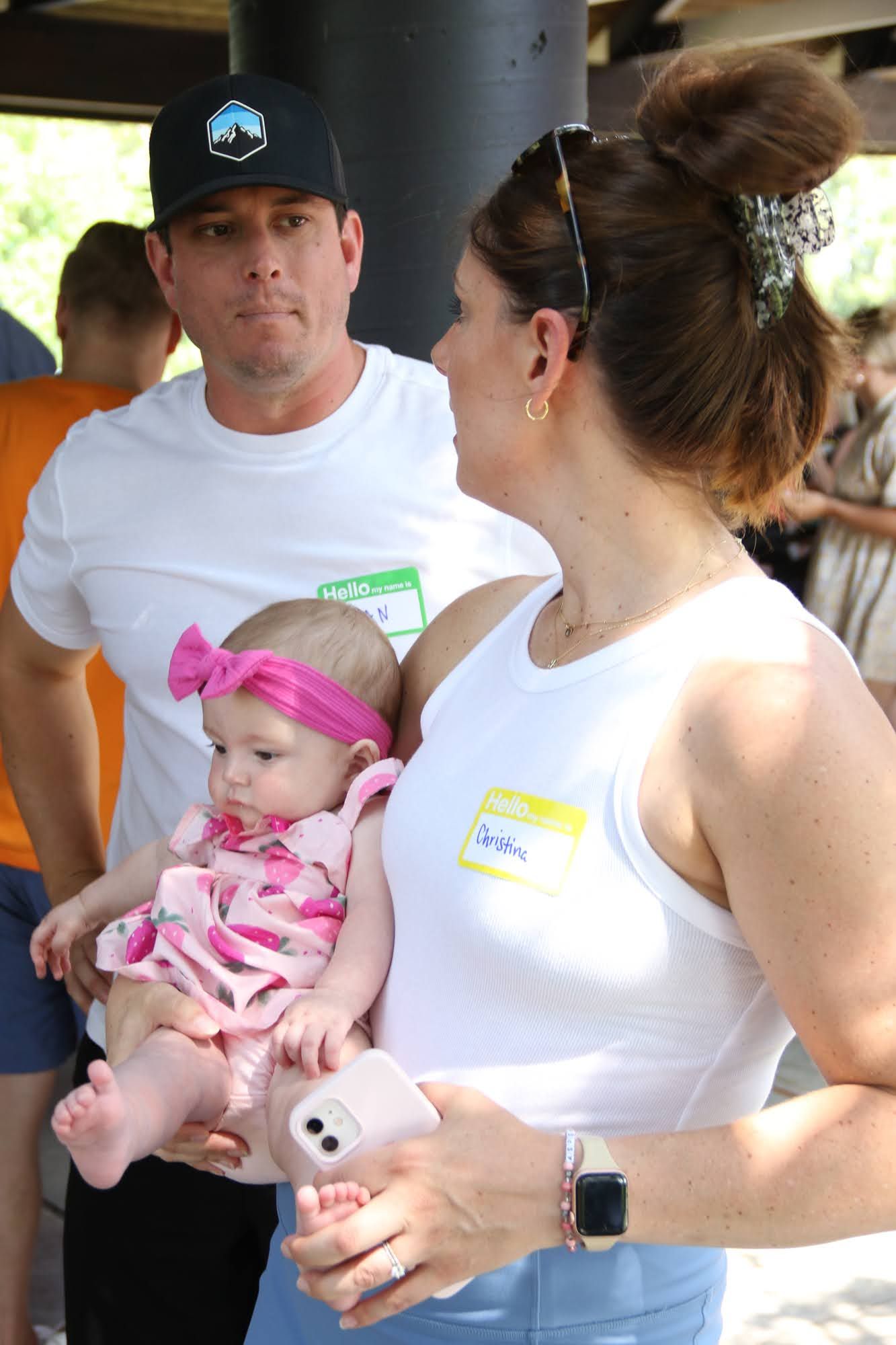 Woman holding baby, man watches; group gathering outside.