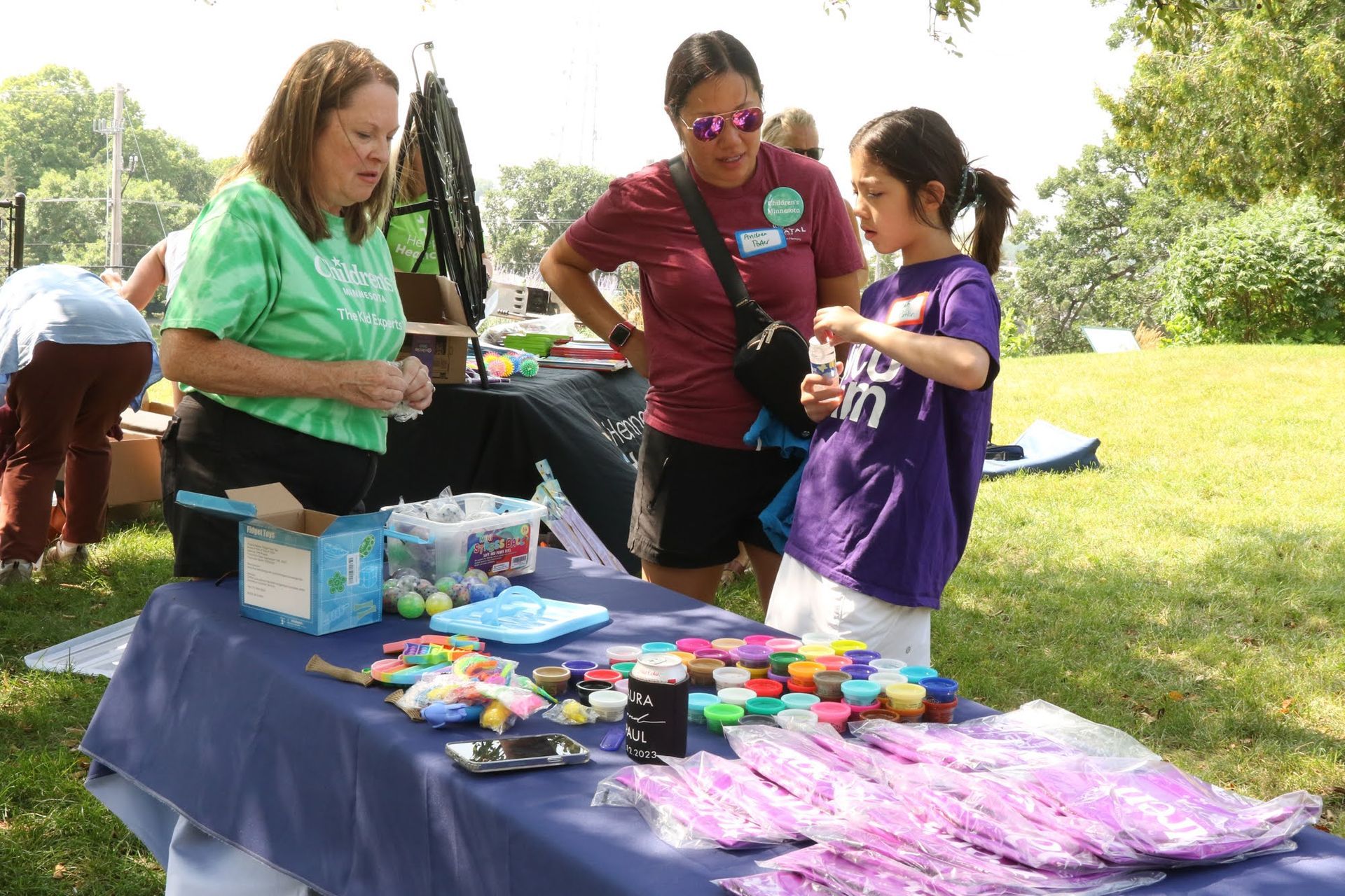 People at a table with items, talking. One person in green shirt, one in red, child in purple. Outdoors, grass.