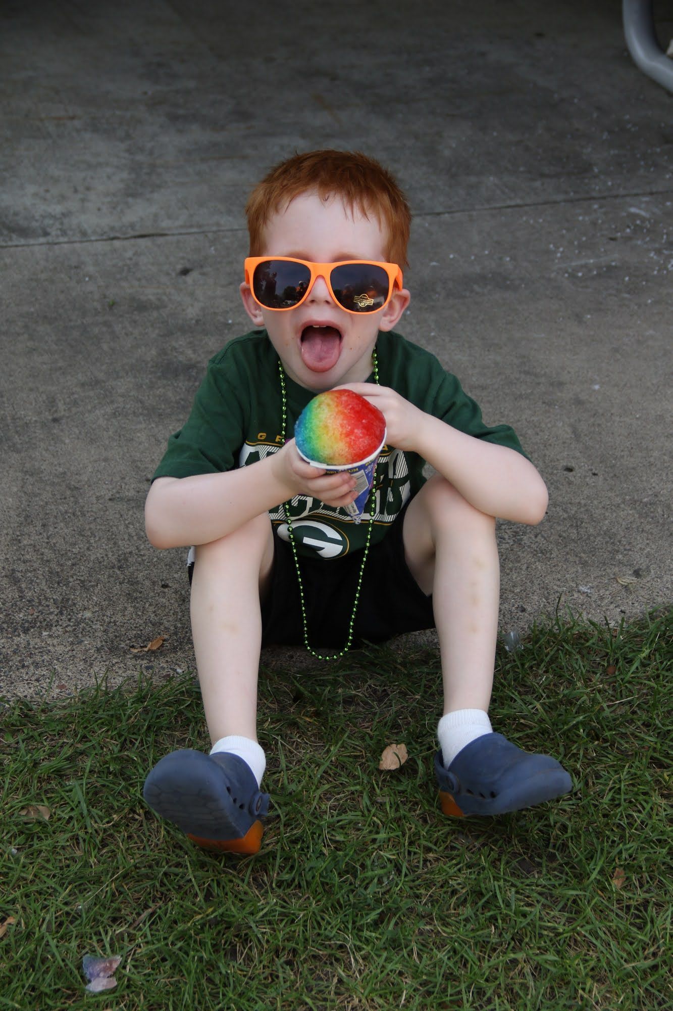 Boy with orange sunglasses and red hair, sitting on grass, holding rainbow-colored treat, mouth open.