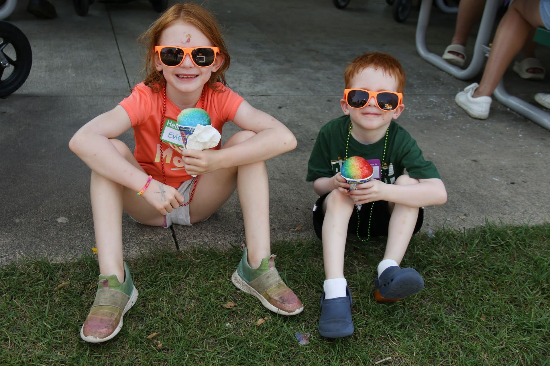 Two children wearing sunglasses, holding snow cones, sitting on grass.