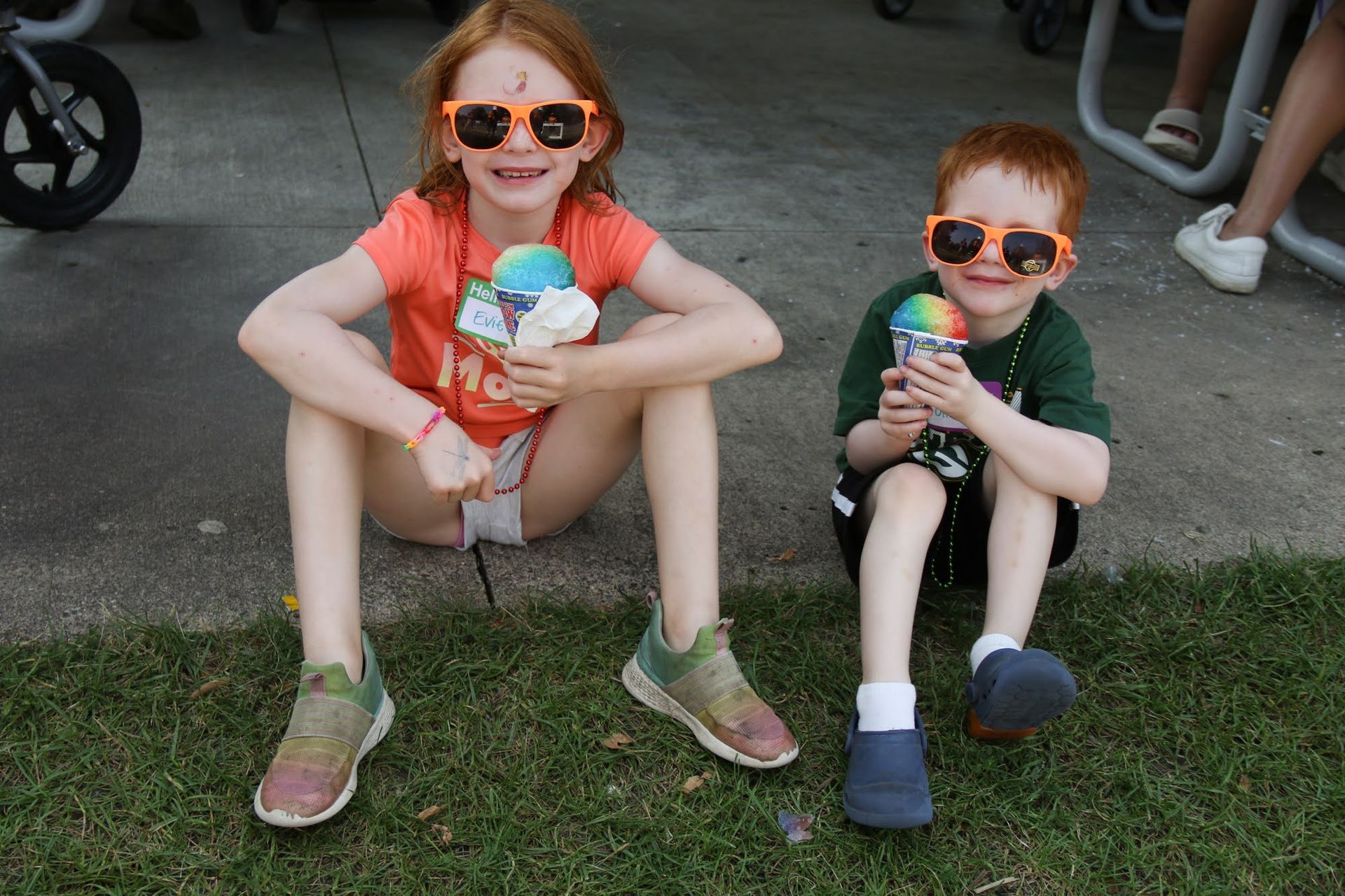 Two children wearing sunglasses enjoy snow cones while sitting outside.