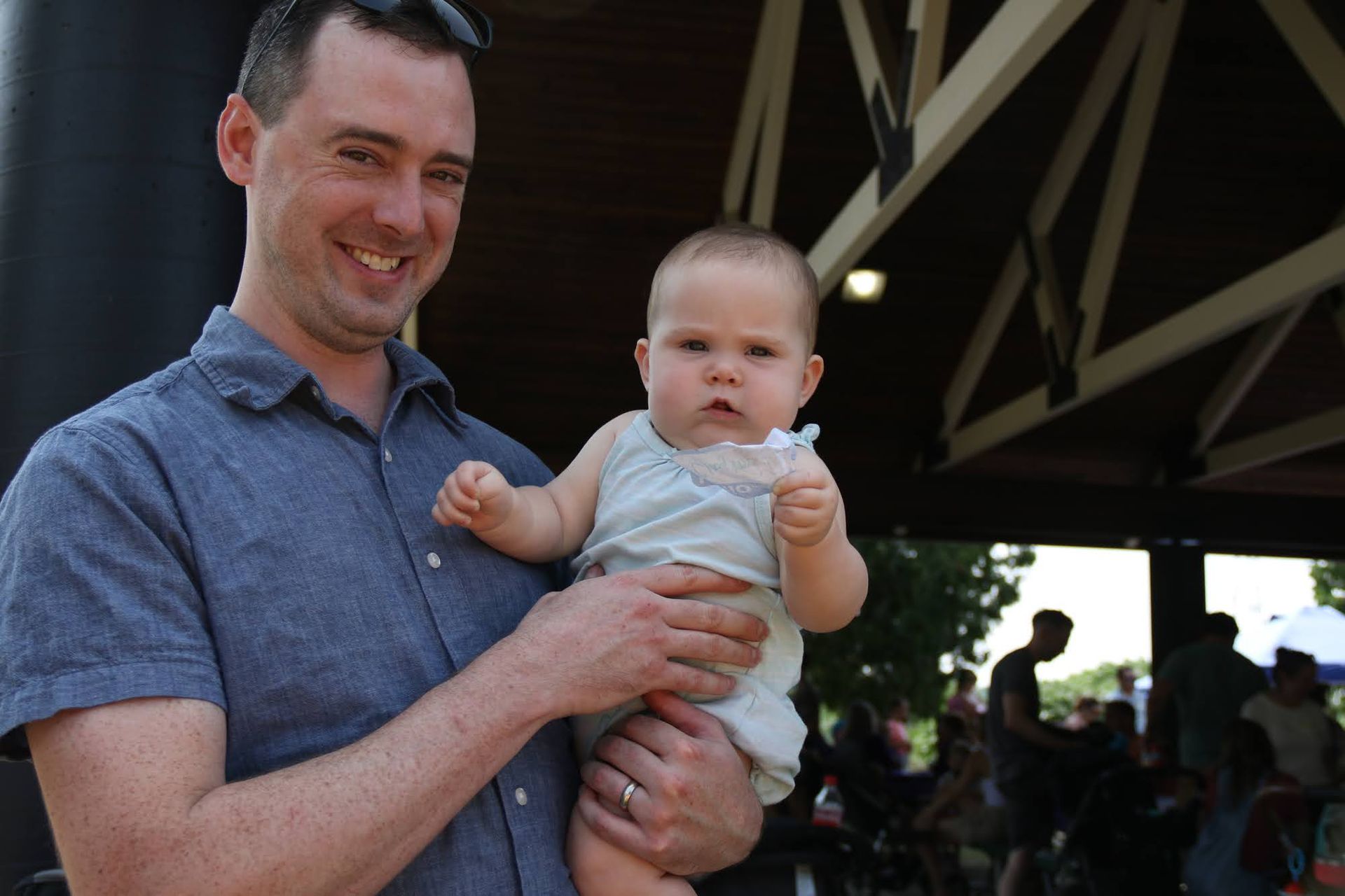 Man holding baby; outdoors. Man smiles; baby has a serious expression, holding a toy.