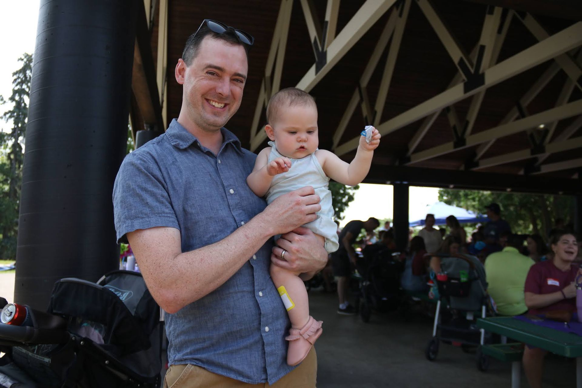 Man holding a baby at an outdoor event under a pavilion. Both are smiling. Other people are in the background.
