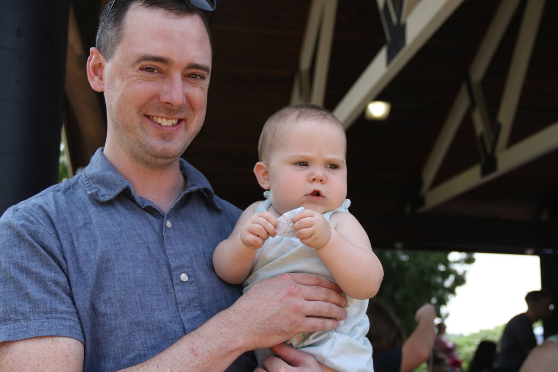 Man holding a baby, both looking at the camera. They are outside under a wooden structure.