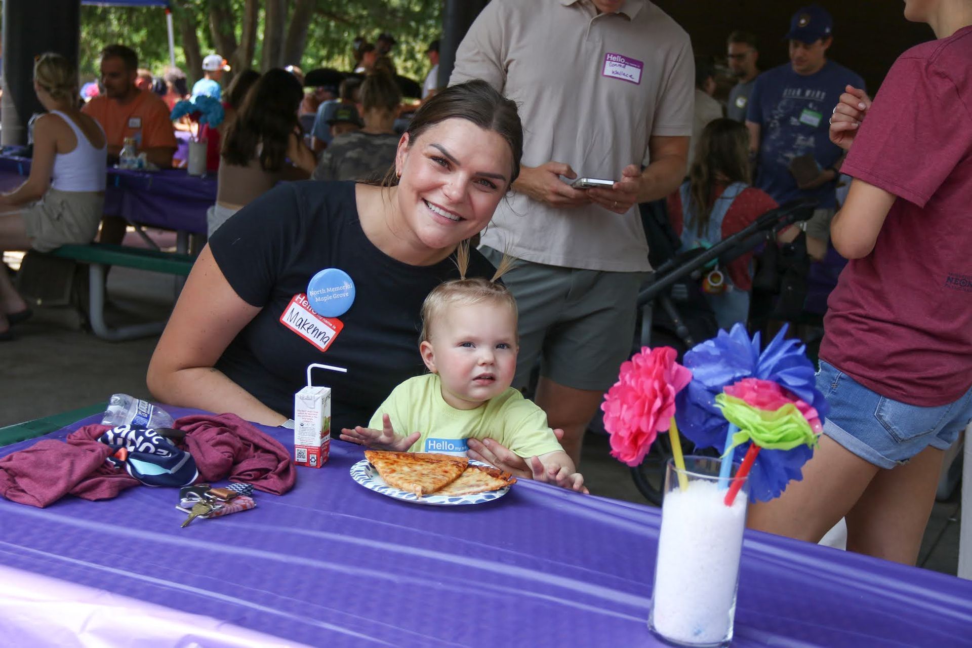 Woman and child at outdoor party, smiling at the camera, purple table, food, drinks, other people in the background.