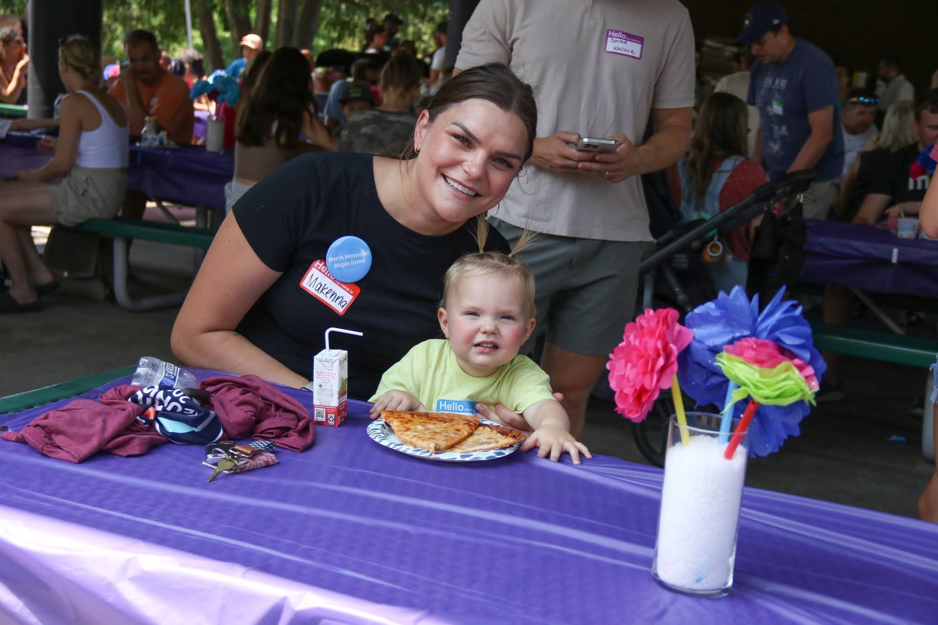 Woman and child at a picnic table, smiling. Purple table cloth, food and drink, colorful paper decorations.