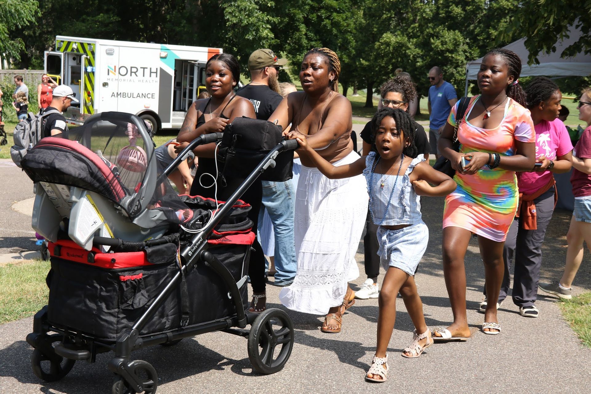 People walking outdoors, some pushing a stroller. A child leads, others follow. Ambulance visible.