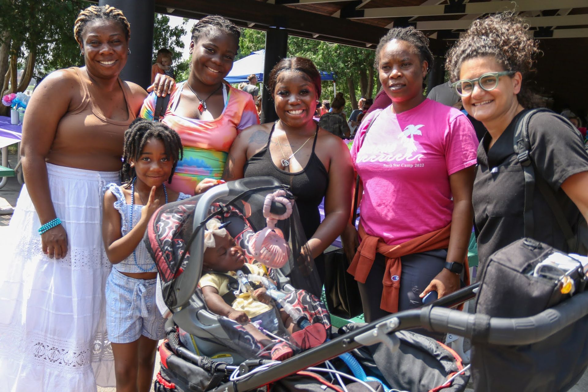 Group of people smiling, posing with a stroller outdoors under a shaded structure.