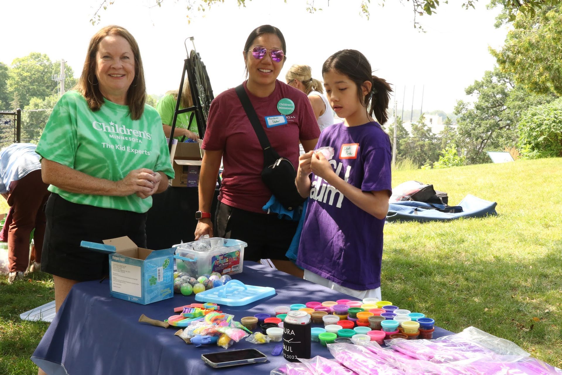 Three people at a table outdoors: one in green shirt, one in burgundy shirt, and a girl in purple shirt, displaying and discussing colorful toys.