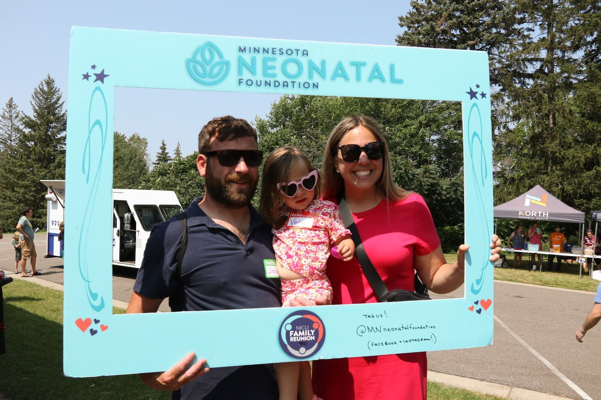 Family poses with a photo frame at an event. A woman, man, and child smile outdoors. The frame is branded with text.
