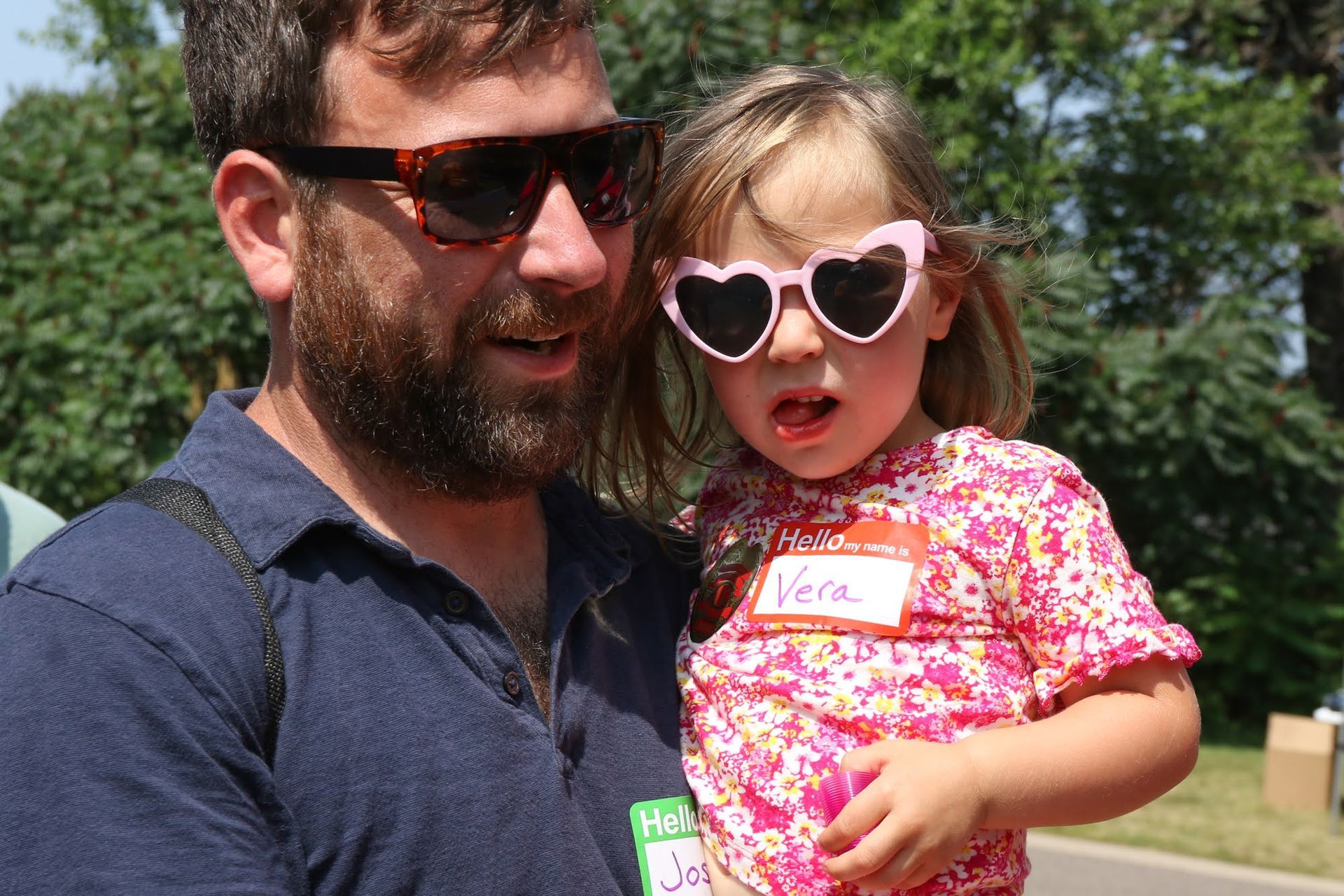 Man holding a child wearing heart-shaped sunglasses. The child has a name tag. Outdoors, sunny day.
