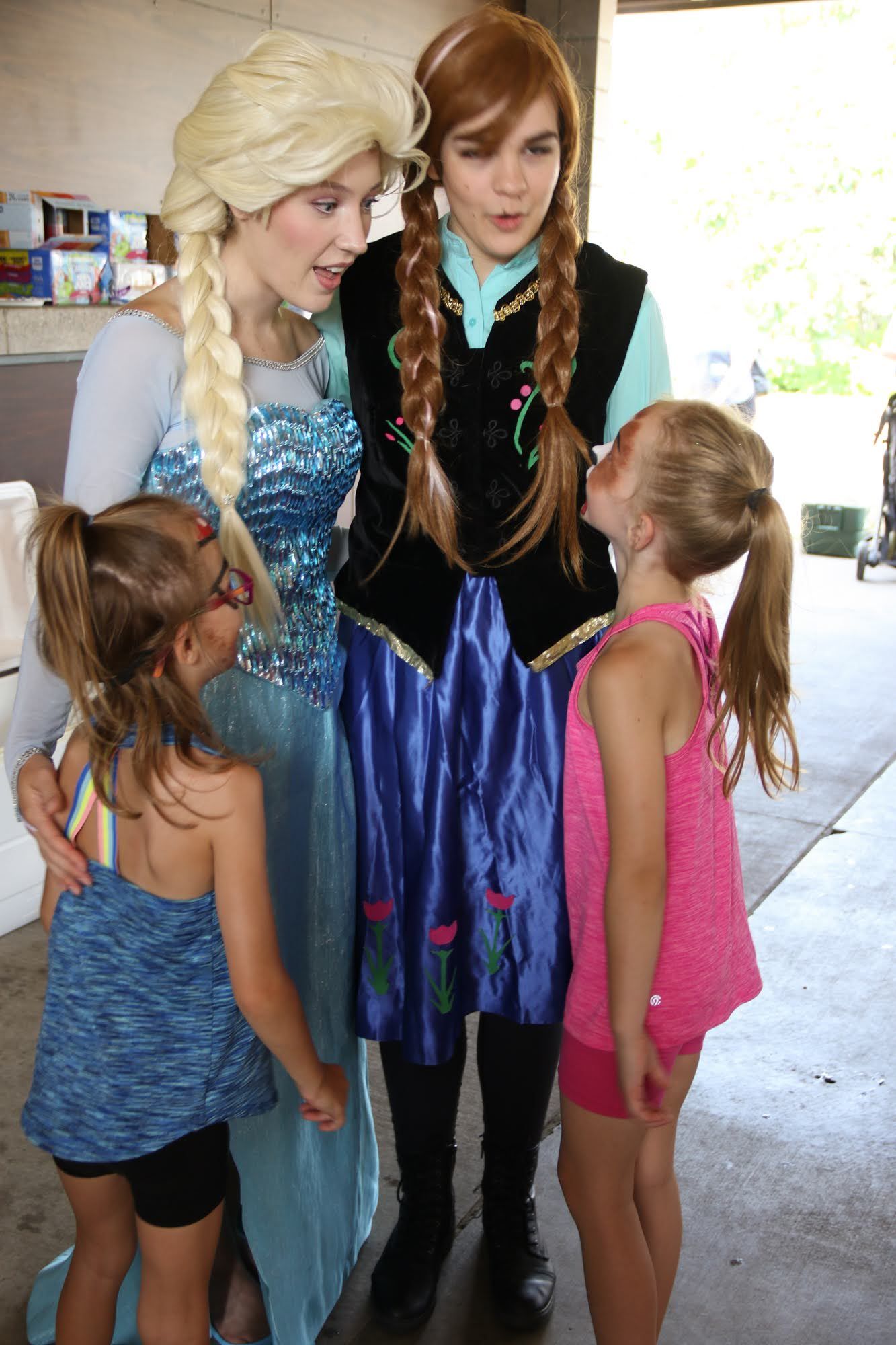 Elsa and Anna posing with two children at an outdoor event. Elsa in a blue dress, Anna in a blue skirt, the kids are in casual wear.