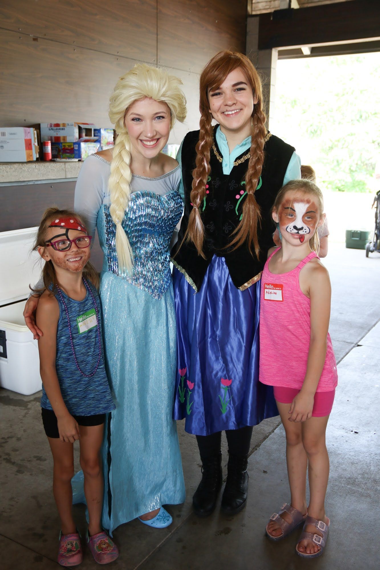 Elsa and Anna costumed characters pose with two children who have face paint at an outdoor event.
