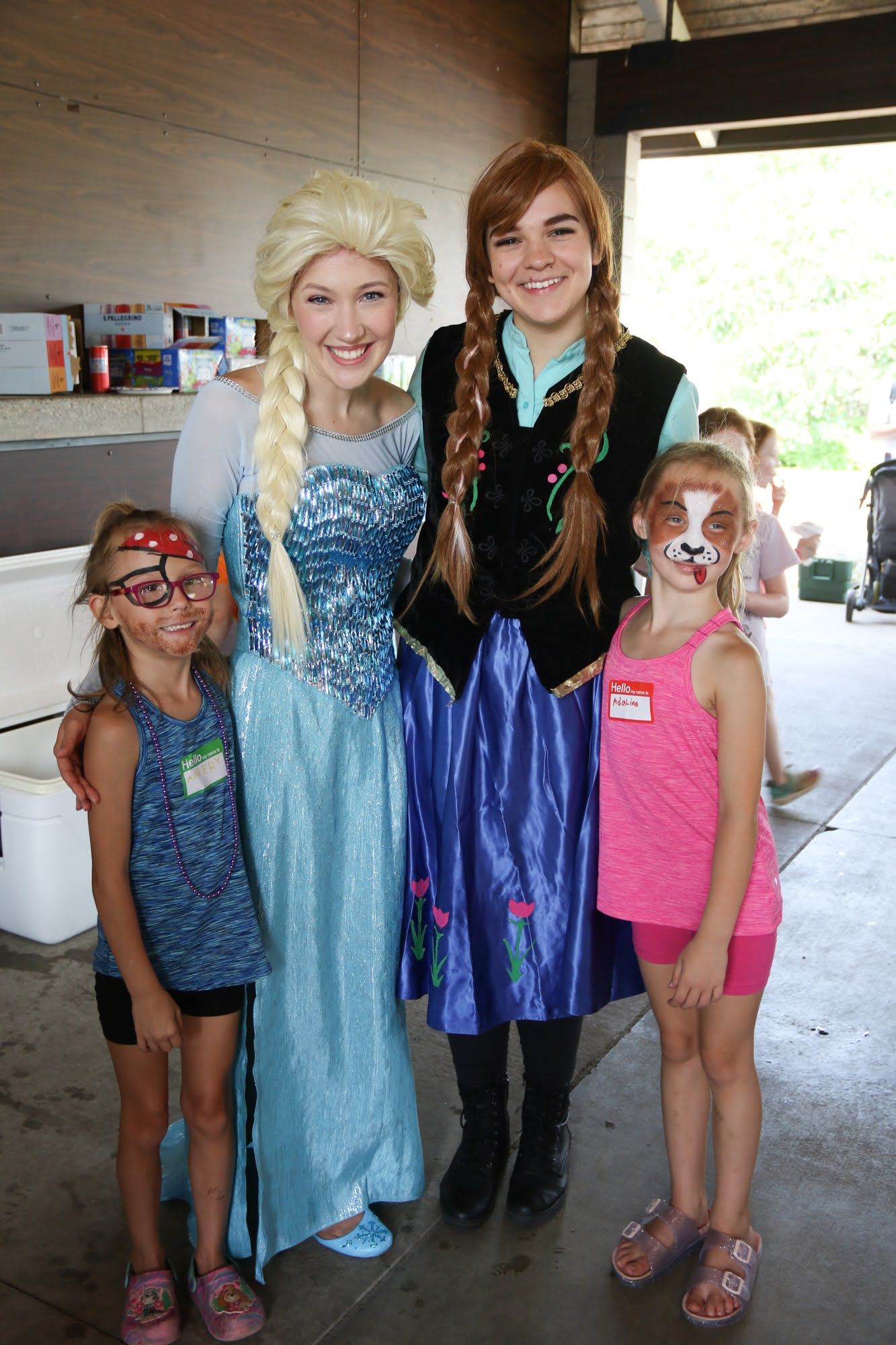Elsa and Anna costumed characters pose with two girls with face paint at an outdoor event.