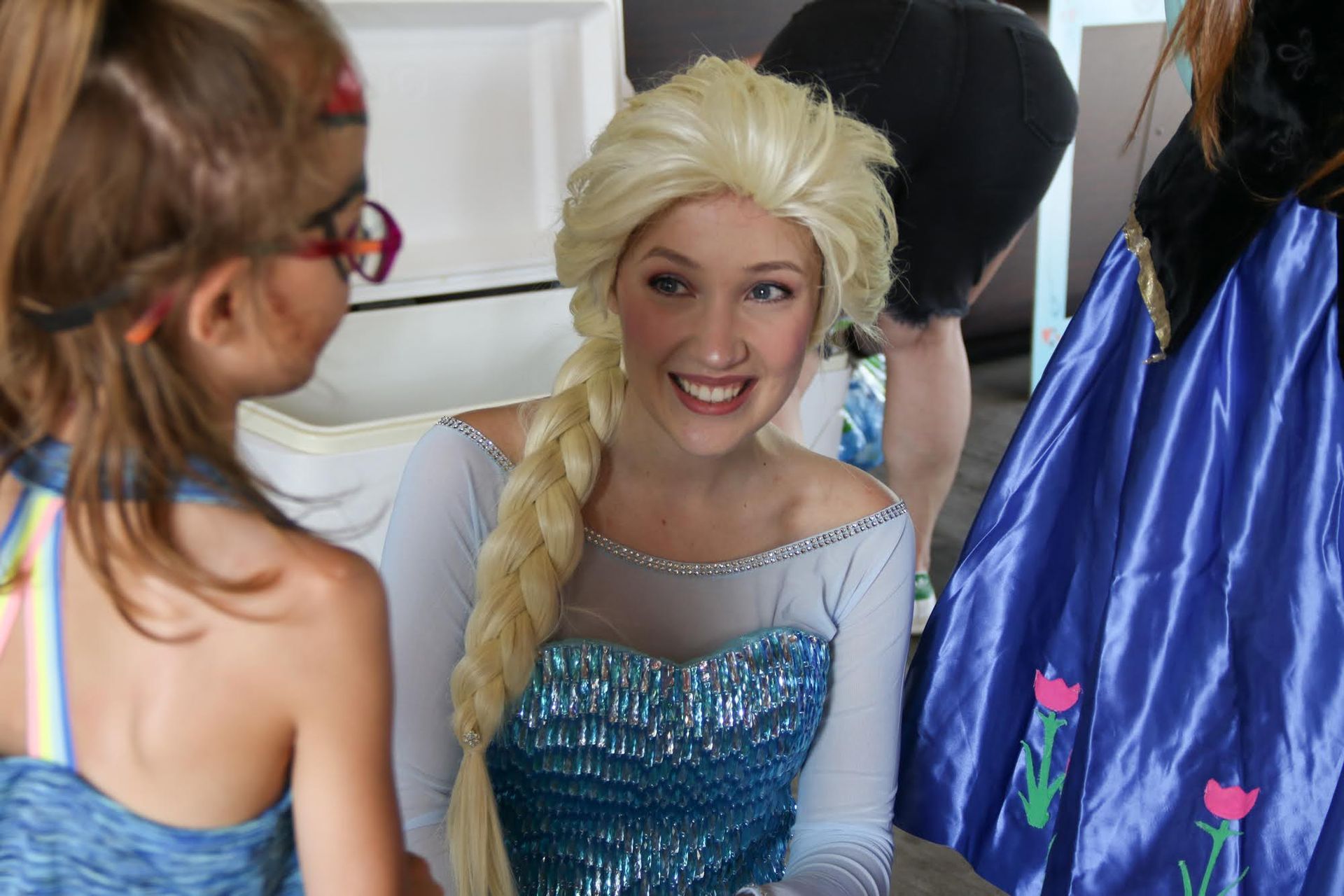 Elsa from Frozen with a child. Elsa wears a blue dress and smiles, talking to the child.