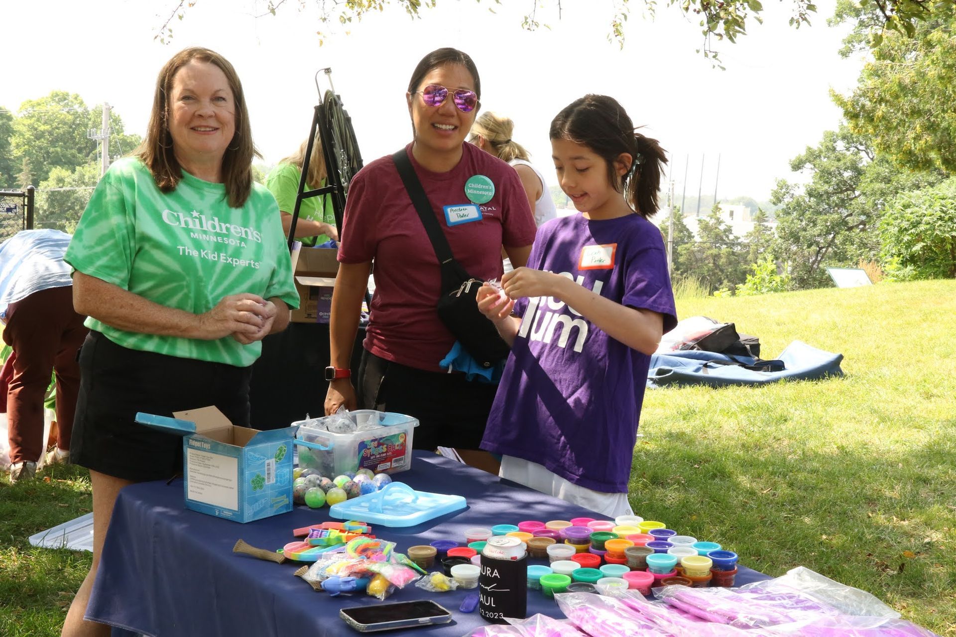 People at a craft table in a park. Woman in green shirt smiles at the camera, girl in purple shirt examines craft items.