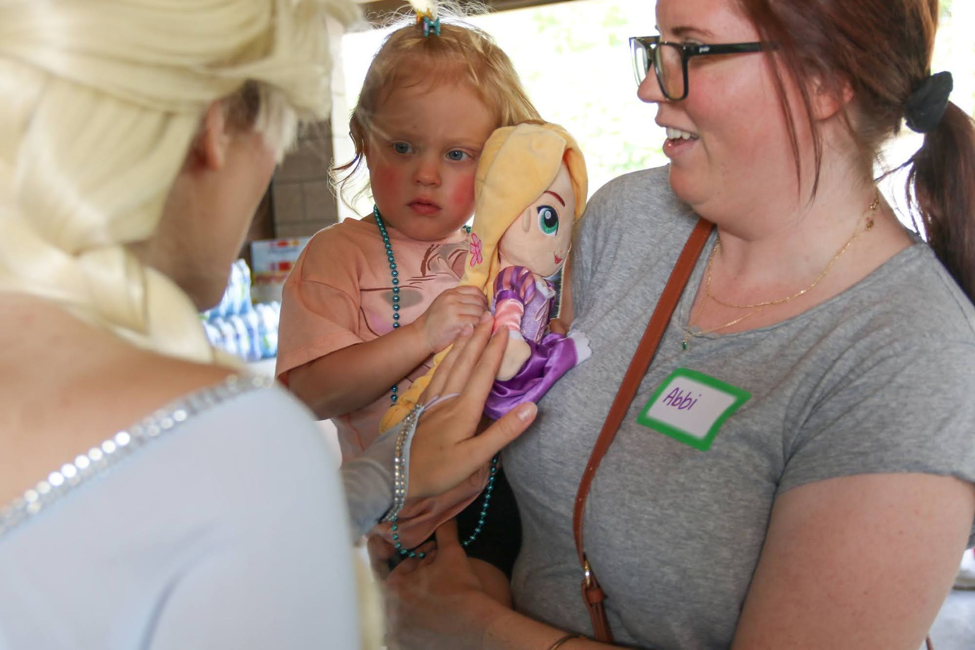 Blond princess talks to a girl holding a doll with an adult. Outdoor setting.