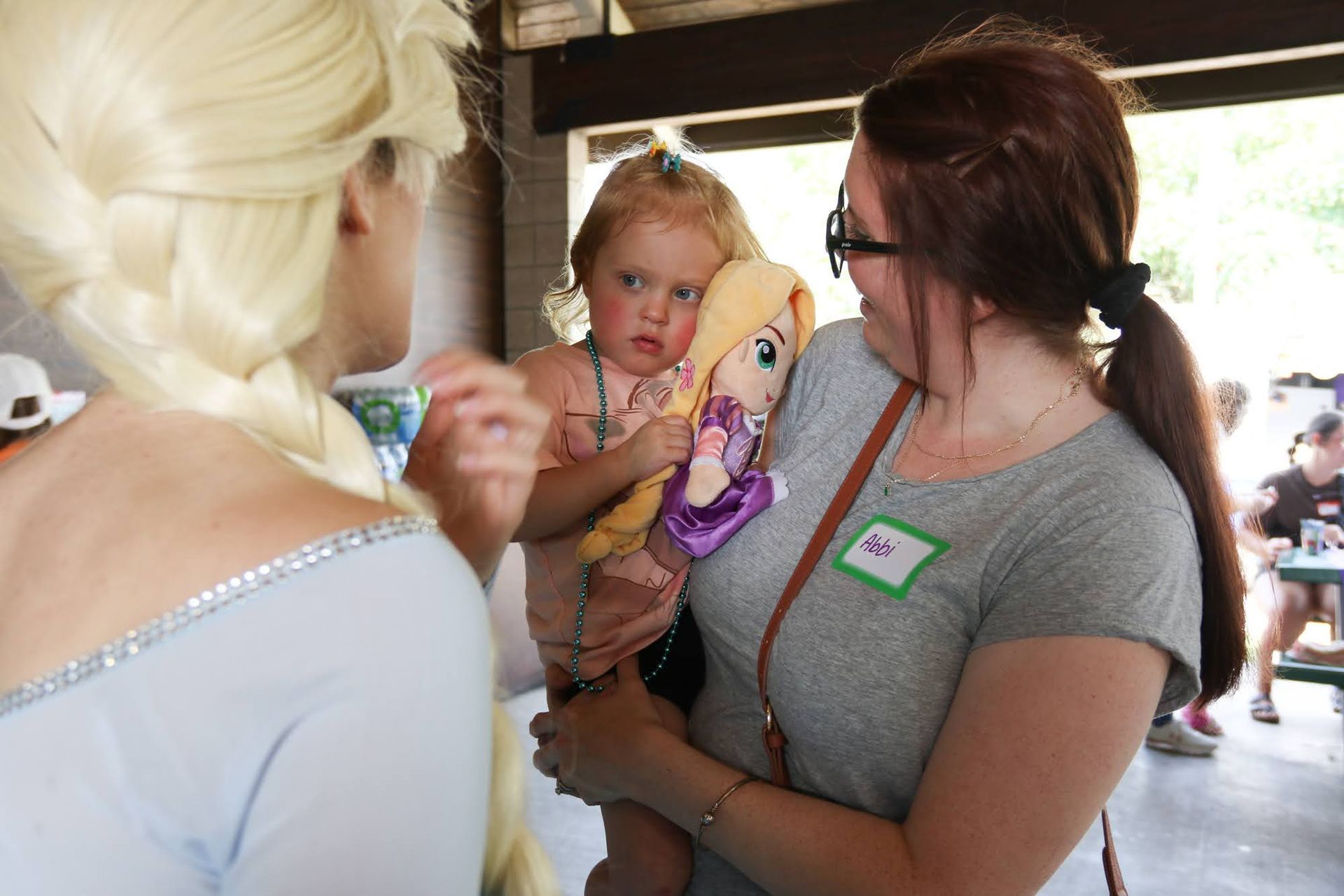 A woman holds a child clutching a doll, talking to someone in an Elsa costume. Outdoors, shelter in background.