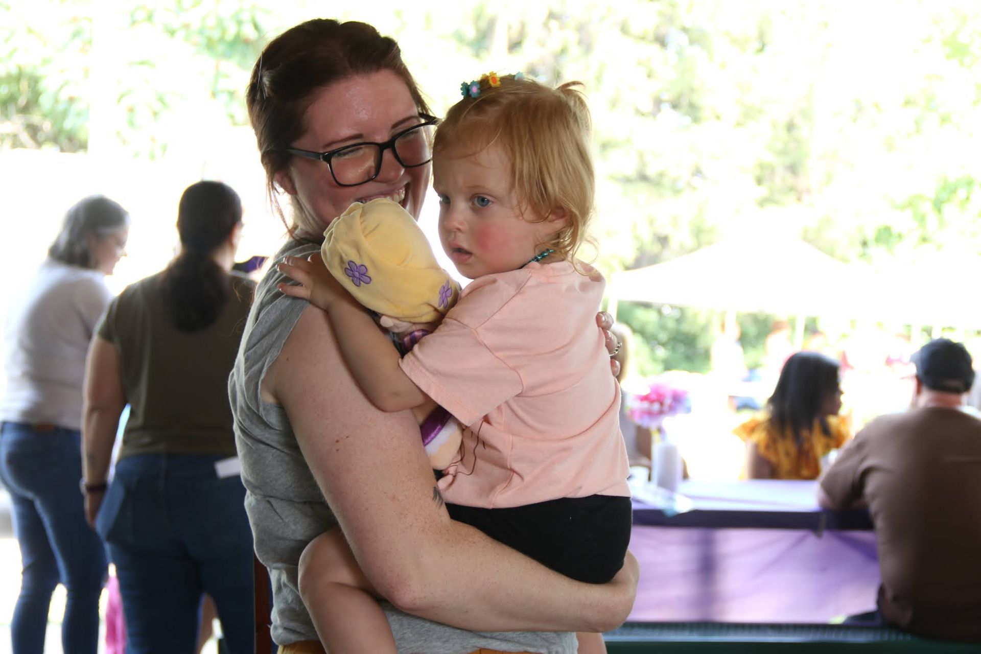 Woman holding a child and a stuffed animal at an outdoor event, people and tables visible in the background.