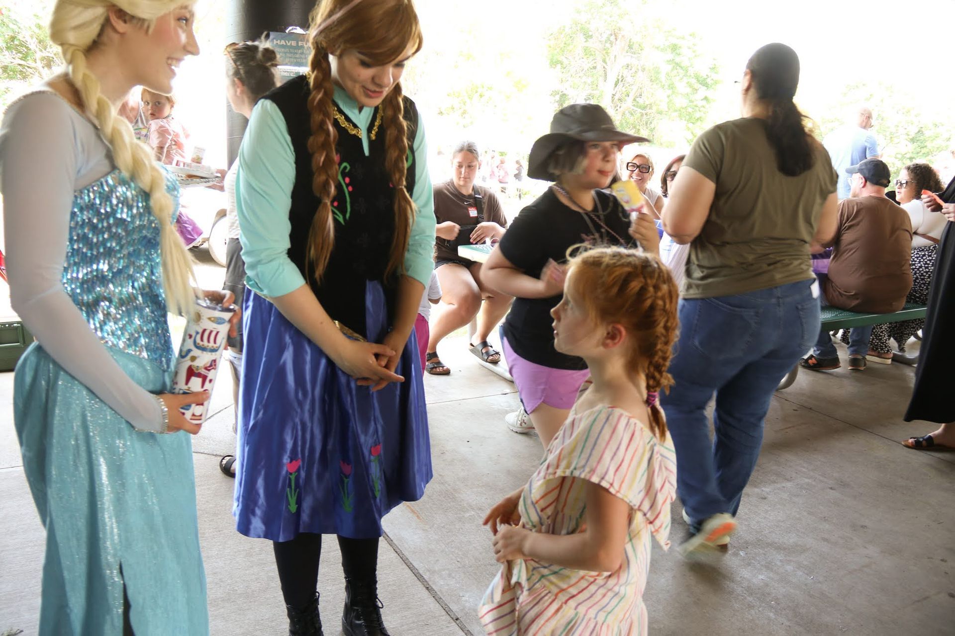 Elsa and Anna costumed characters with a child at an outdoor event. Others watch.