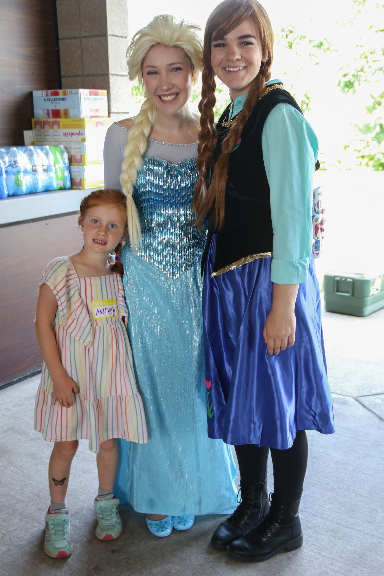 Elsa and Anna characters pose with a young girl. They are outside. The girl wears a colorful dress.