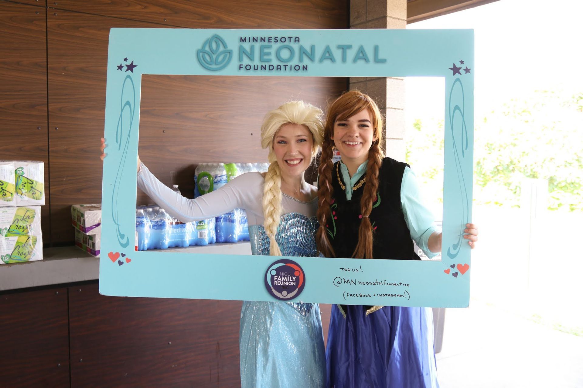 Elsa and Anna posing with a frame at a Minnesota Neonatal Foundation event; both smiling.