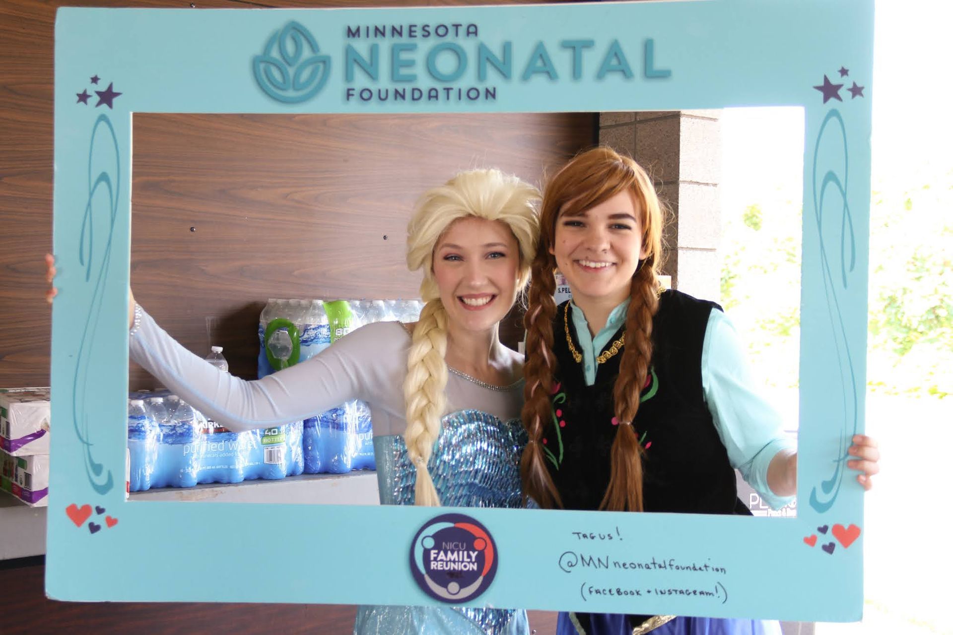 Two people dressed as Elsa and Anna pose behind a blue photo frame for the Minnesota Neonatal Foundation.