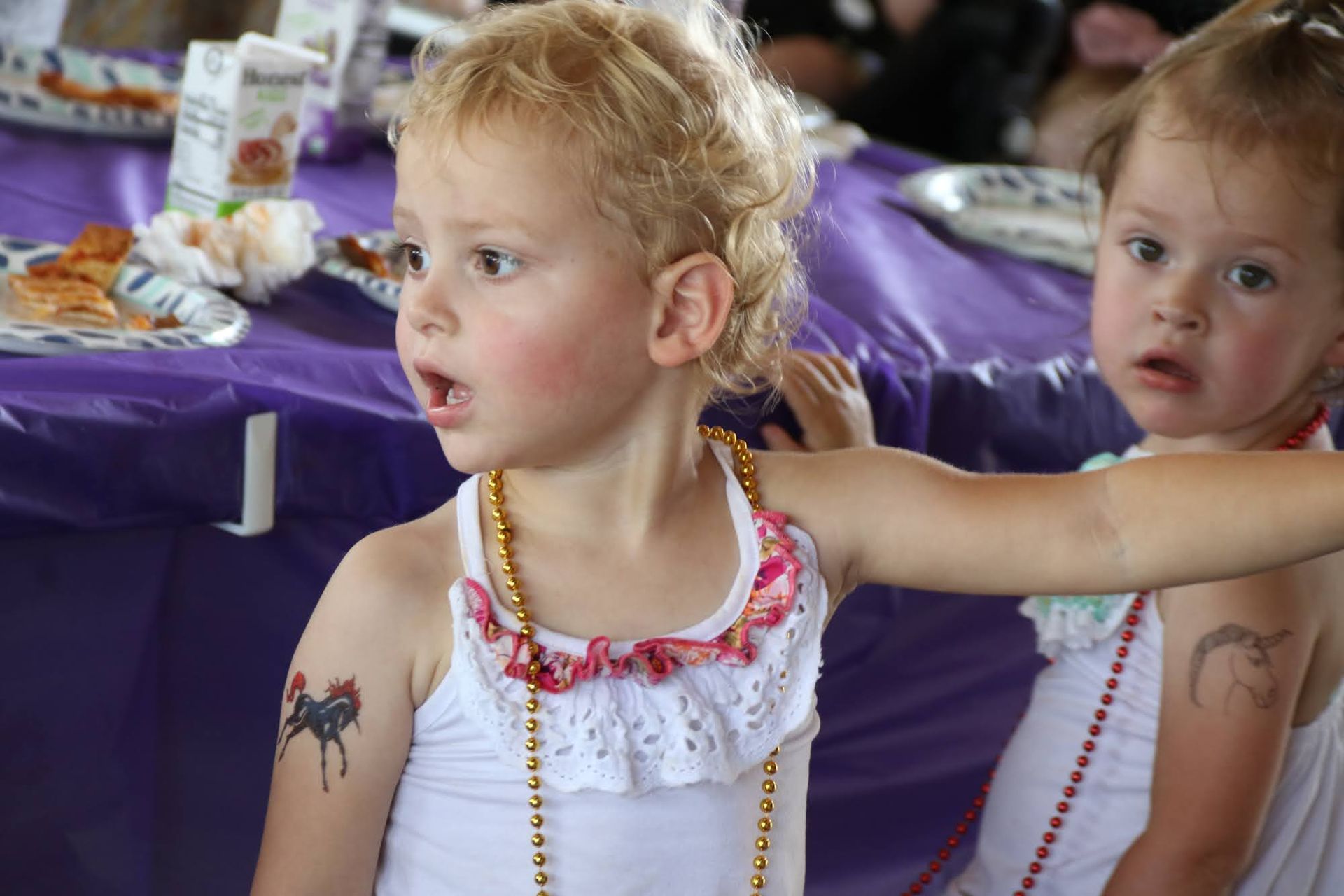 Two young children at a table, one with a shocked expression and a tattoo, wearing beads.