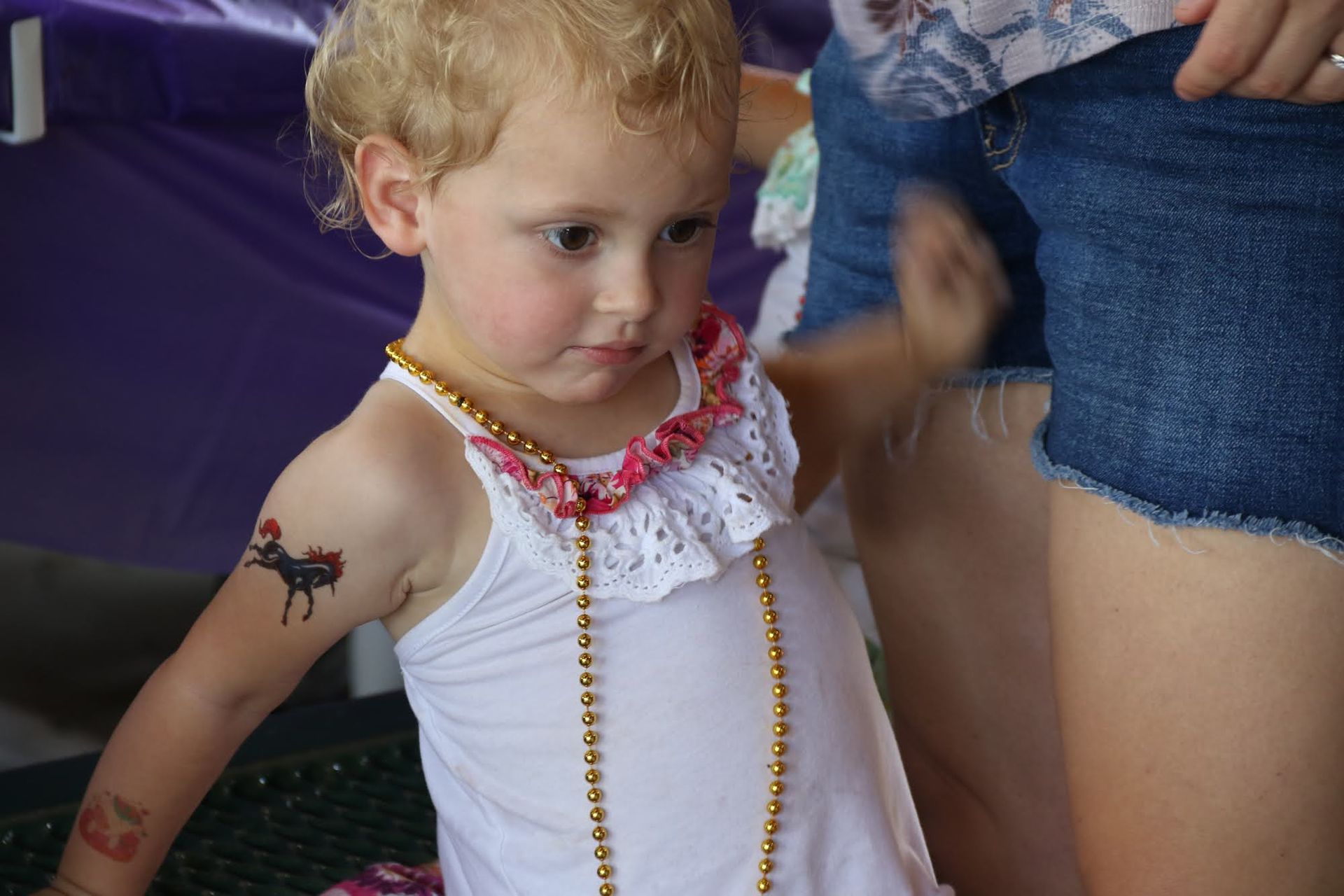 Blond child in white tank top with necklace and temporary arm tattoos, looking down.