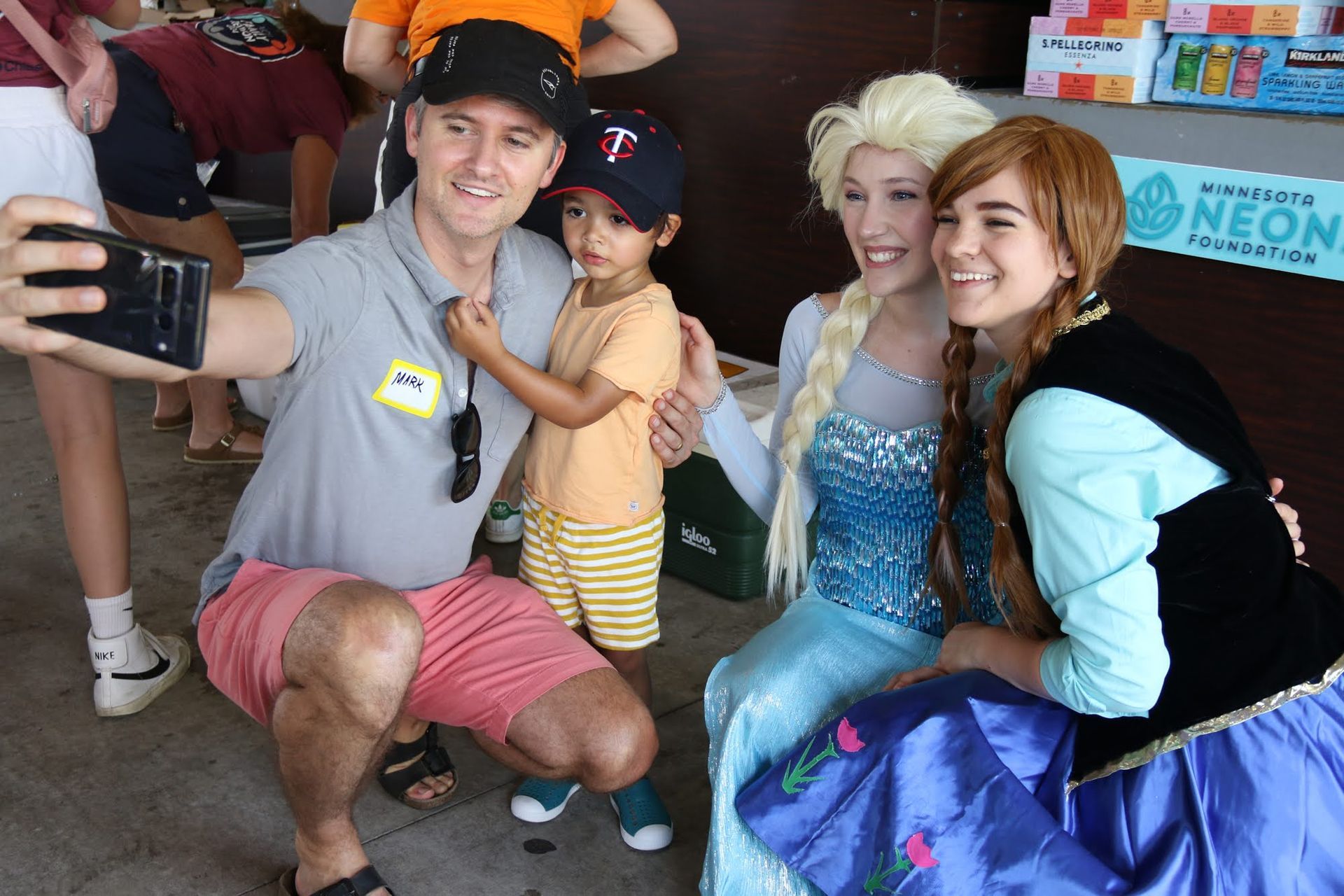 Man taking selfie with a child and two costumed princesses at an outdoor event.