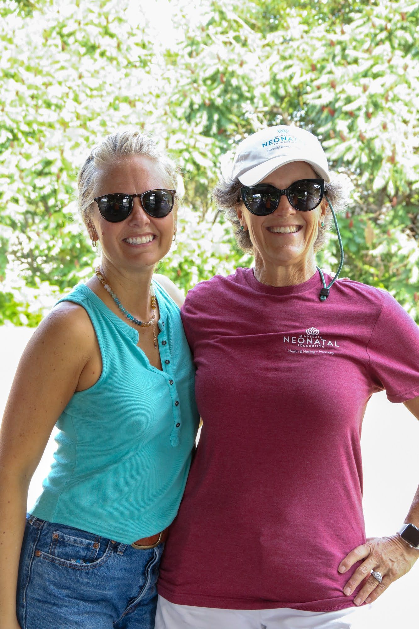 Two women in sunglasses smile; one in a turquoise tank top and denim, the other in a maroon t-shirt and white cap.