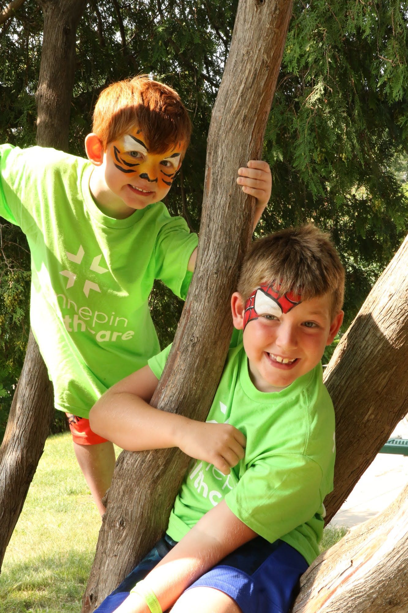 Two children with face paint climb a tree, wearing green shirts and shorts, smiling.
