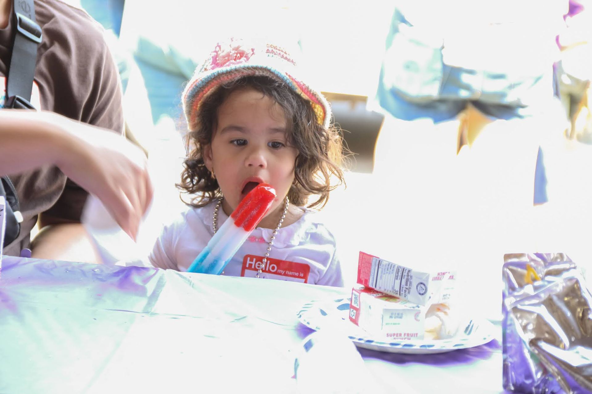 Child eating a popsicle at an outdoor event. They wear a hat and a name tag that says 