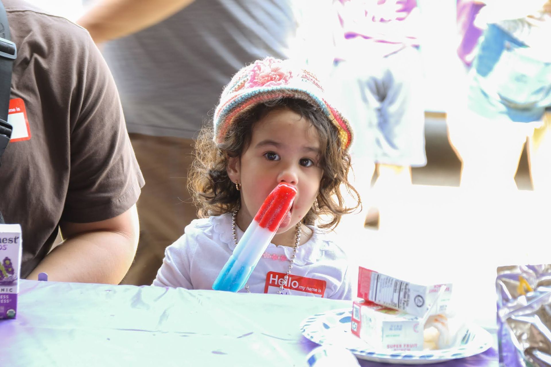 Child eating a popsicle at a table; wears a hat and has a name tag.