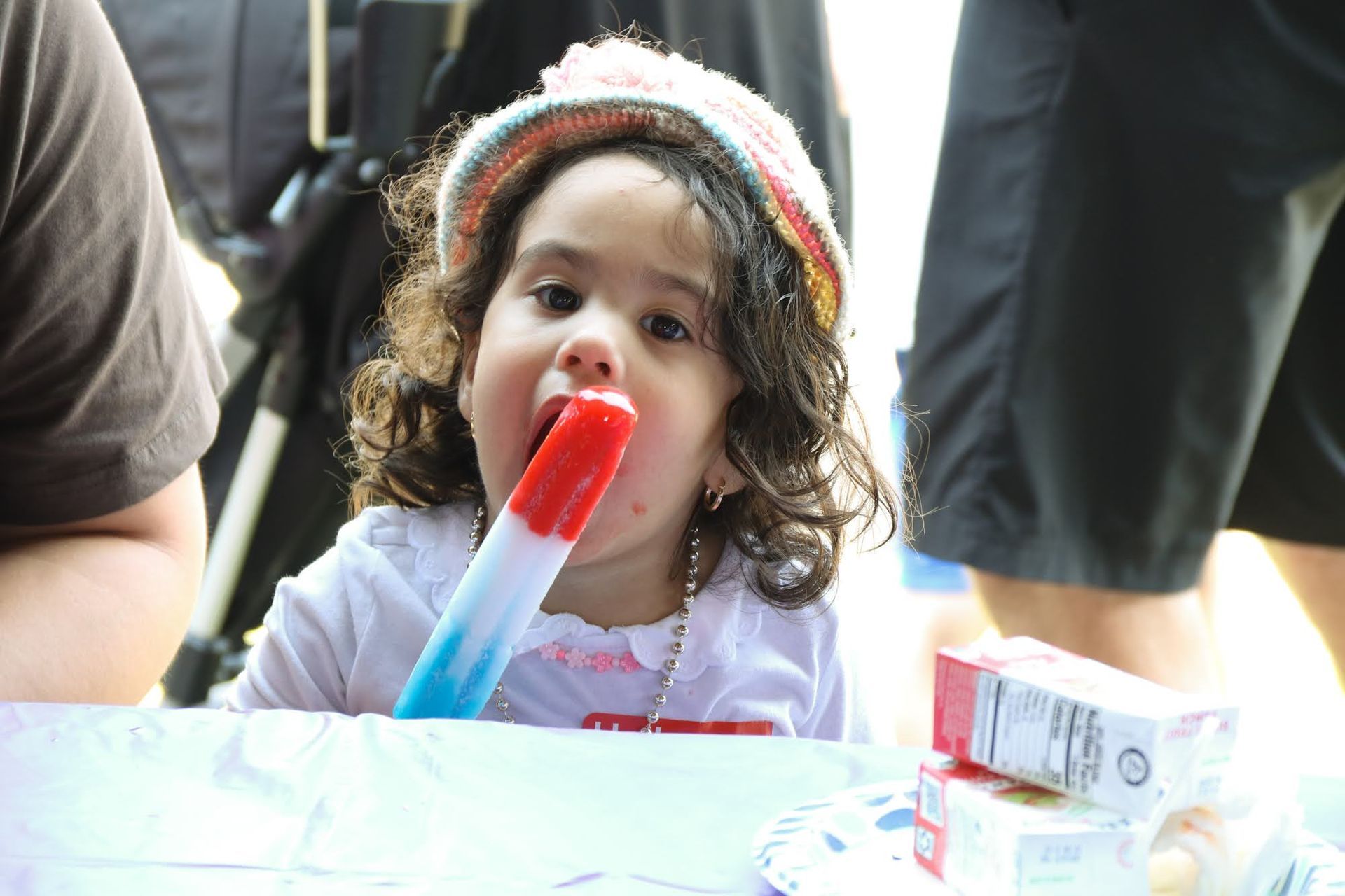 Child eating a red, white, and blue popsicle outdoors, wearing a hat, smiling at the camera.