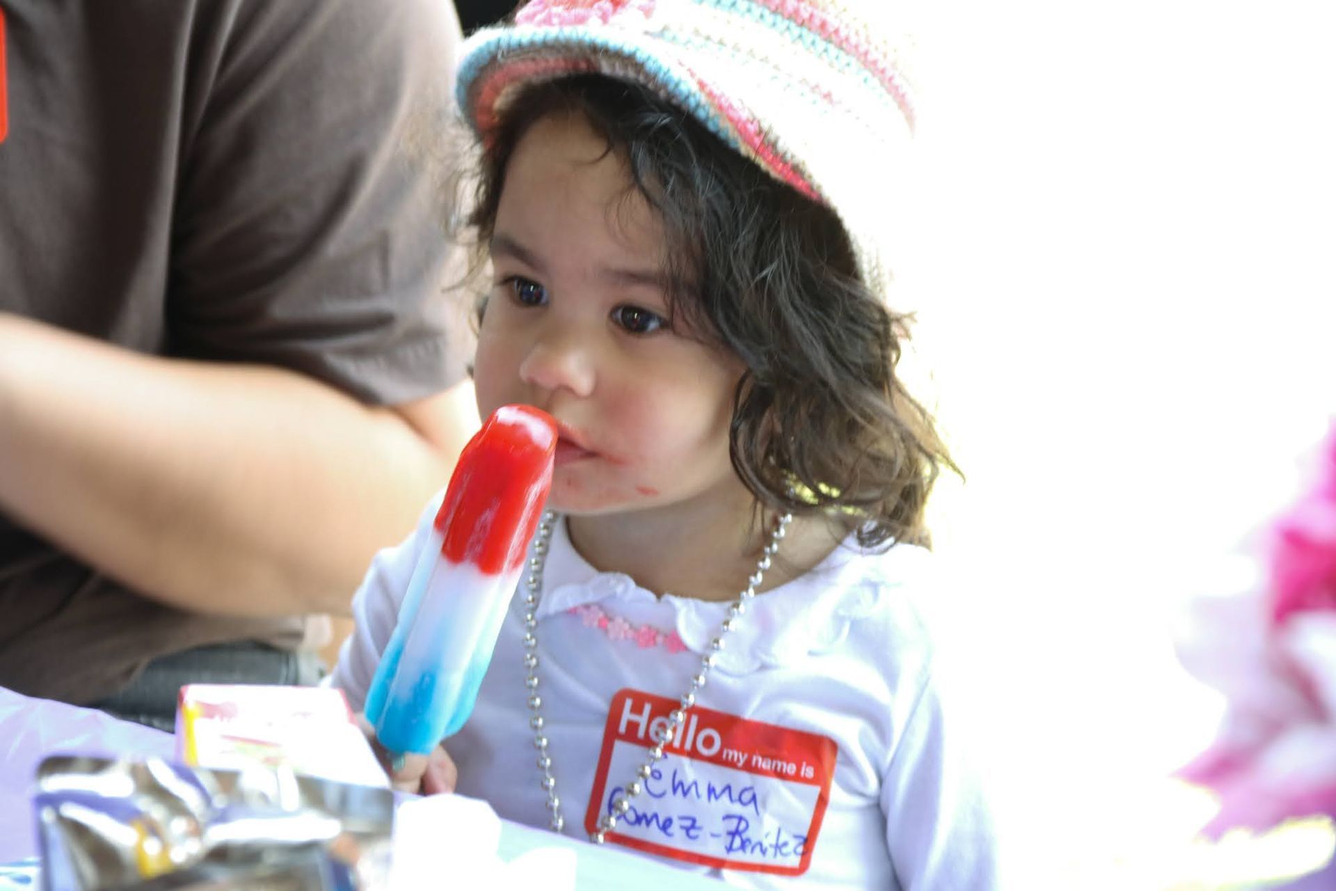 Girl eating a red, white, and blue popsicle, wearing a hat and name tag.