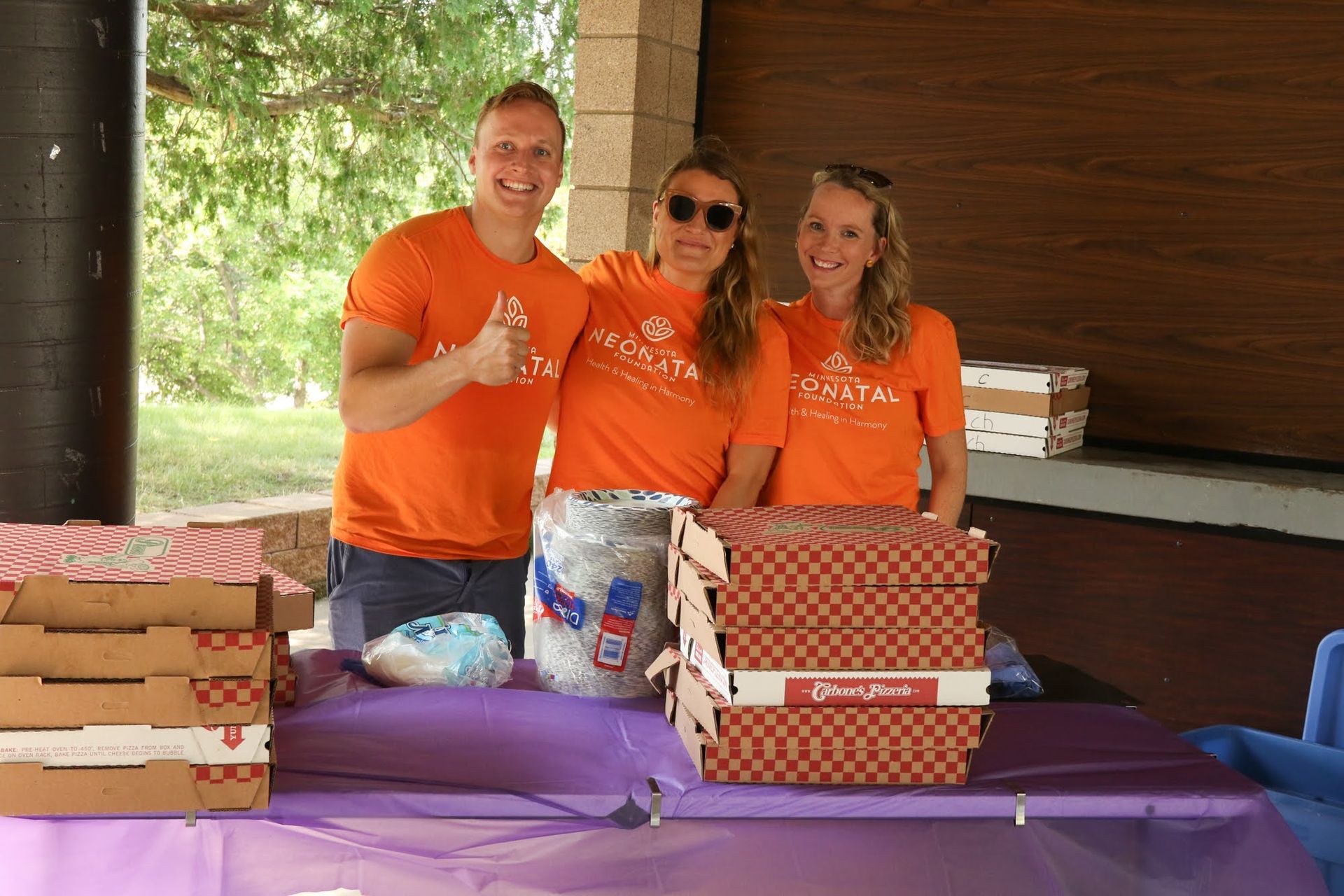 Three people in orange shirts stand behind a table with pizza boxes.