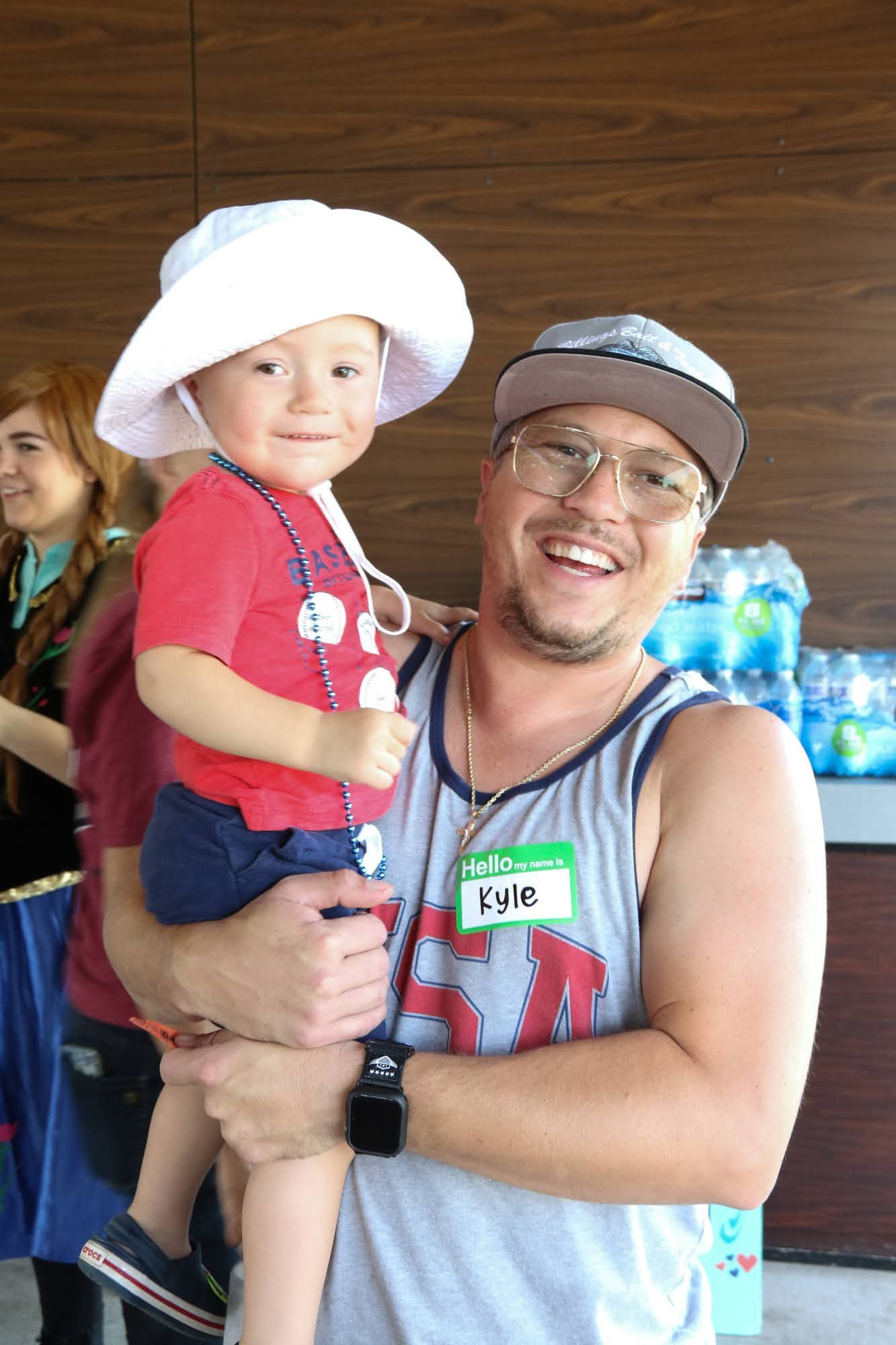 Man holding a child wearing a hat, smiling. Background: water bottles, another person in costume.