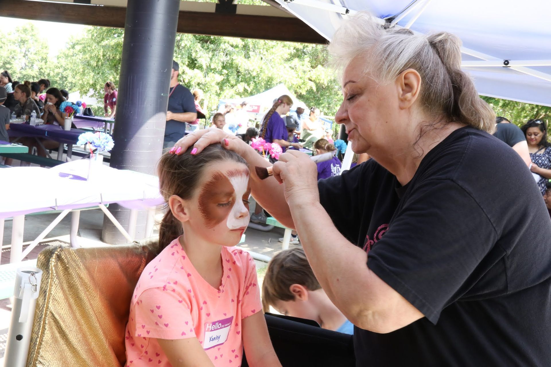 Woman applying face paint to a child at an outdoor event.